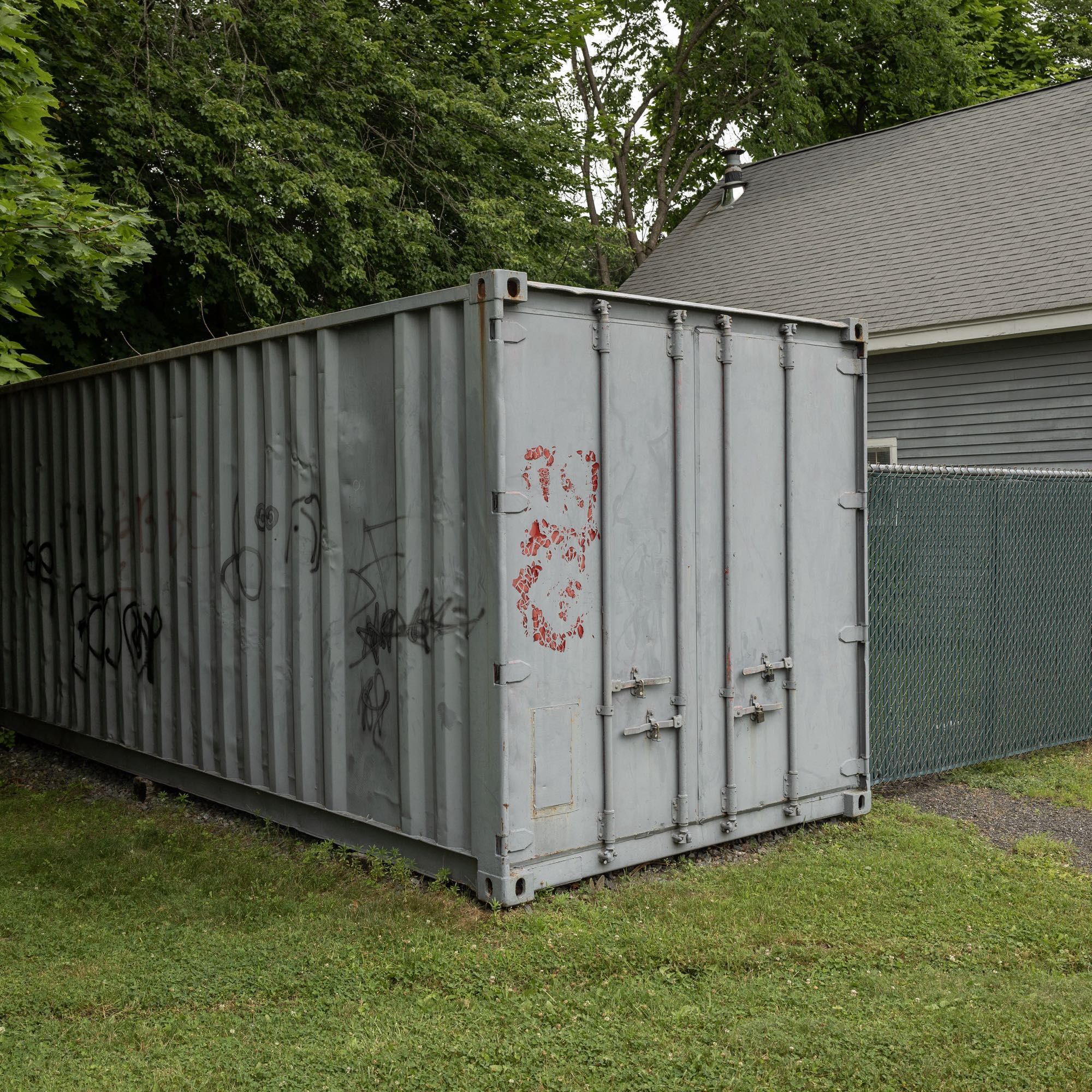 Color photo of a grey shipping container that has some simple graffiti painted on it, photographed with one corner facing the camera. To the right, flush with the face of the container, is a cyclone dense with green privacy material attached to it. Beyond, on the right, is a grey clapboard building with a grey roof. Foreground is grass, background (area above the manmade elements) are leafy trees nearly blocking out the whole hazy sky.