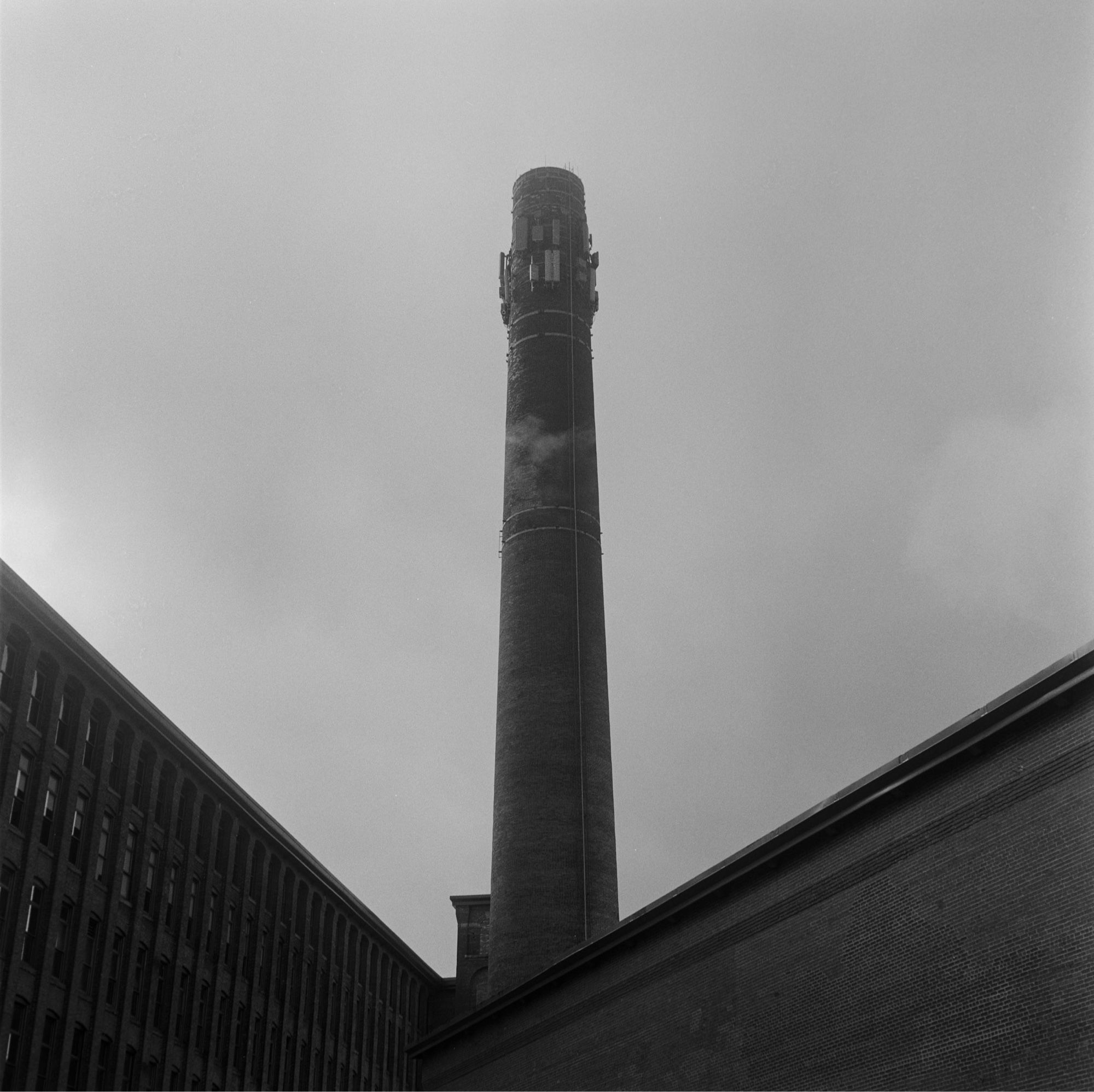 Black and white photo of the smokestack of an old, brick mill complex. Two buildings of the mill meet at almost a 90 degree angle, their rooflines forming a “V” that meets in the lower center of the image almost exactly where the smokestack rises up into a hazy, cloudy sky. A wisp of a cloud passes in front of part of the smokestack.