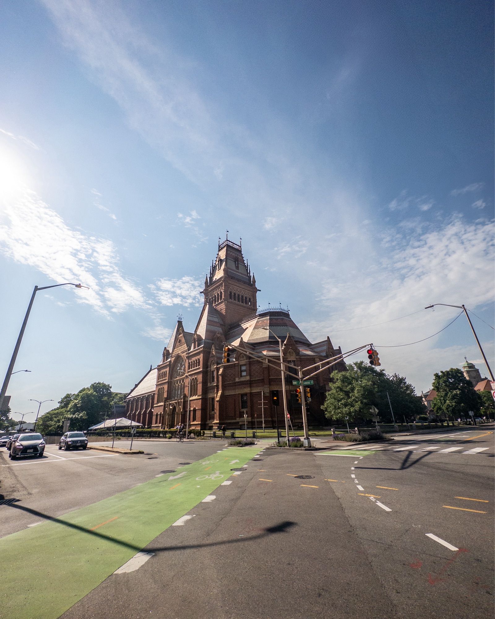 Color photo of Harvard’s Sanders Theatre/Annenberg Hall from the southwest corner of the intersection of Quincy Street and Cambridge Street. A large brick/stone structure of gothic design, the photo depicts it in a vertical composition with a fair amount of blue sky above (a few wispy clouds too) and the heavily marked (bike lanes, travel lines) pavement below. The scene is also peppered with street lamps, trees, some cars near the intersection. One street sign that partially bisected the sky was removed with A.I.