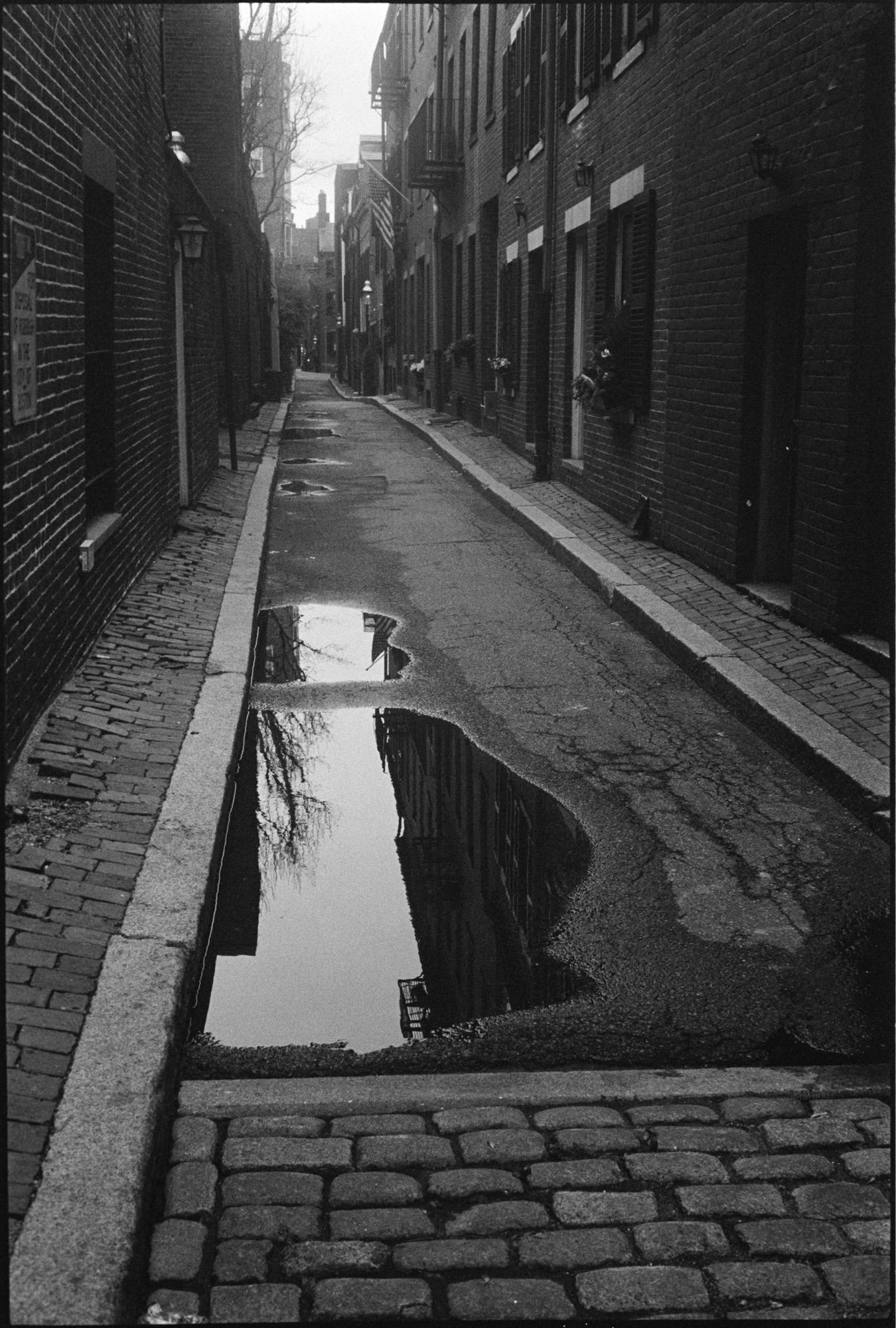 Black and white vertical photo of a narrow alley between a series of brick buildings in Boston’s Beacon Hill neighborhood. Some cobblestones are visible in the foreground but the remainder of the alley is paved with asphalt. As the scene recedes into the distance, an American flag can be seen hanging from one building and the atmosphere becomes increasingly hazy, giving the flavor of 19th century London.