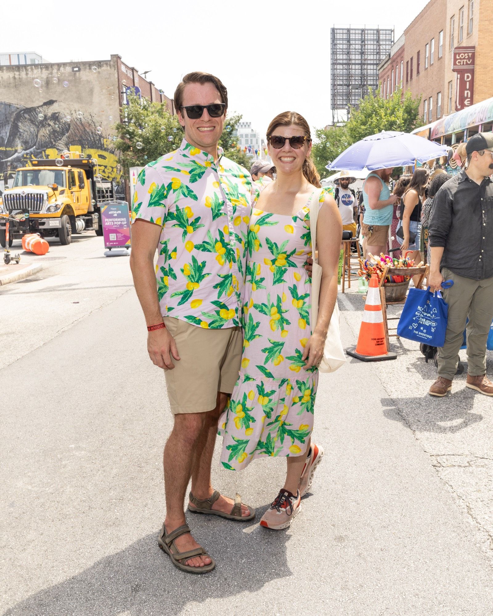 full length street portrait of a smiling man and smiling woman. He is wearing sunglasses, khaki shorts, and a short sleeved shirt with lemons and leaves on it. She is wearing sunglasses and a long sleeveless dress with the same exact lemon and leaf motif. Background is a street fair with pedestrians, parasols, exhibit tents, and buildings.