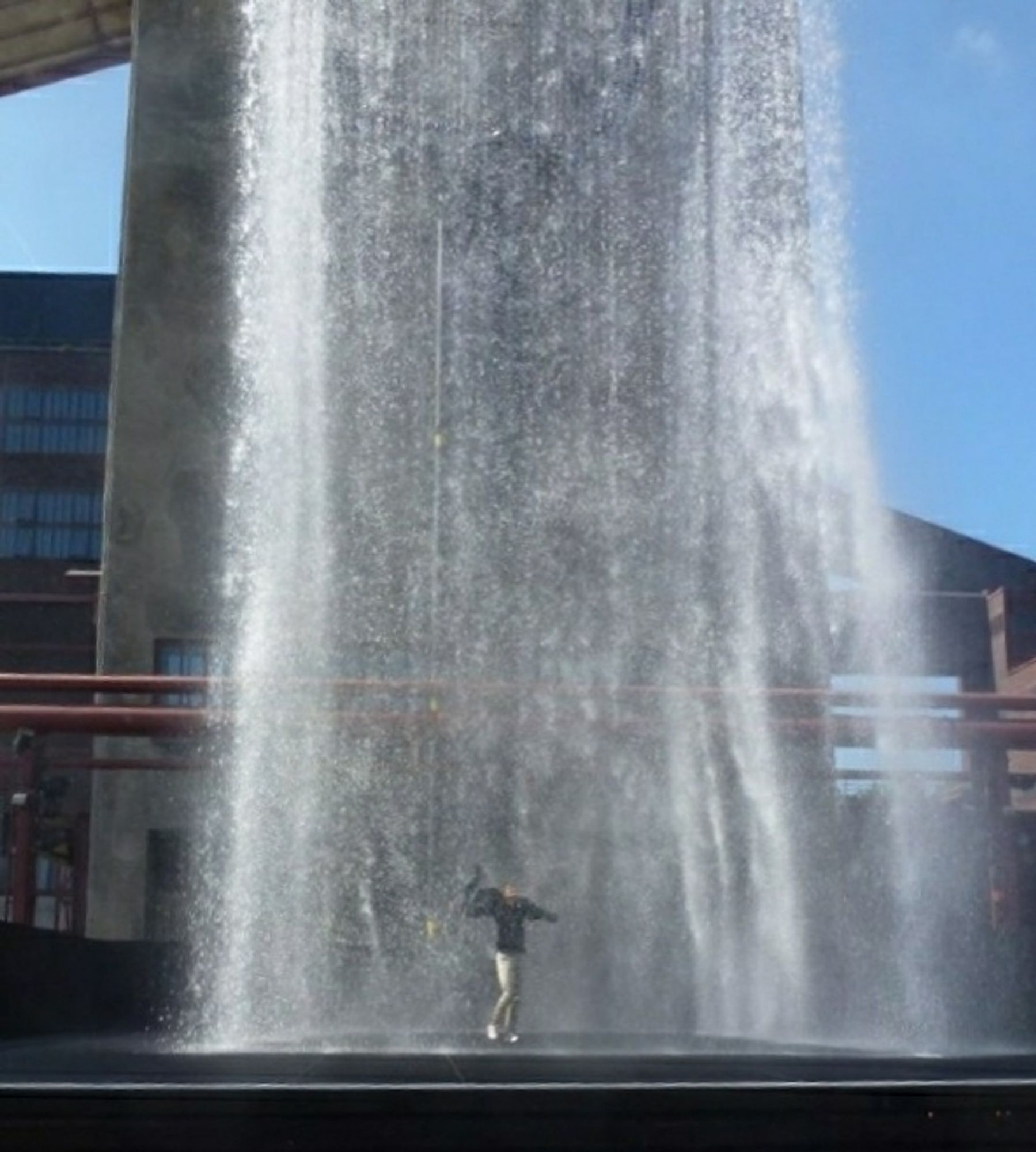 Wasserskulptur "Tower": ein 19 Meter hoher Turm aus Wassertropfen. Hinter dem Wasservorhang steht eine Frau in wehendem, dunkelem Regenschutz und heller Hose und schaut nach oben.