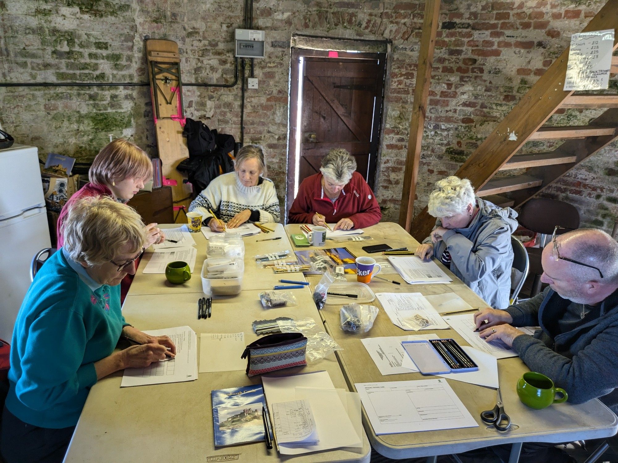 Six people sat around a table drawing. Brick curved wall in background.