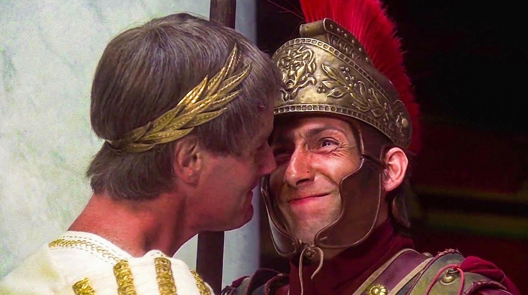 a shot from the “biggus dickus” scene from Monty Python’s Life of Brian. two men are really close together, one confronting the other, both trying to stifle laughter