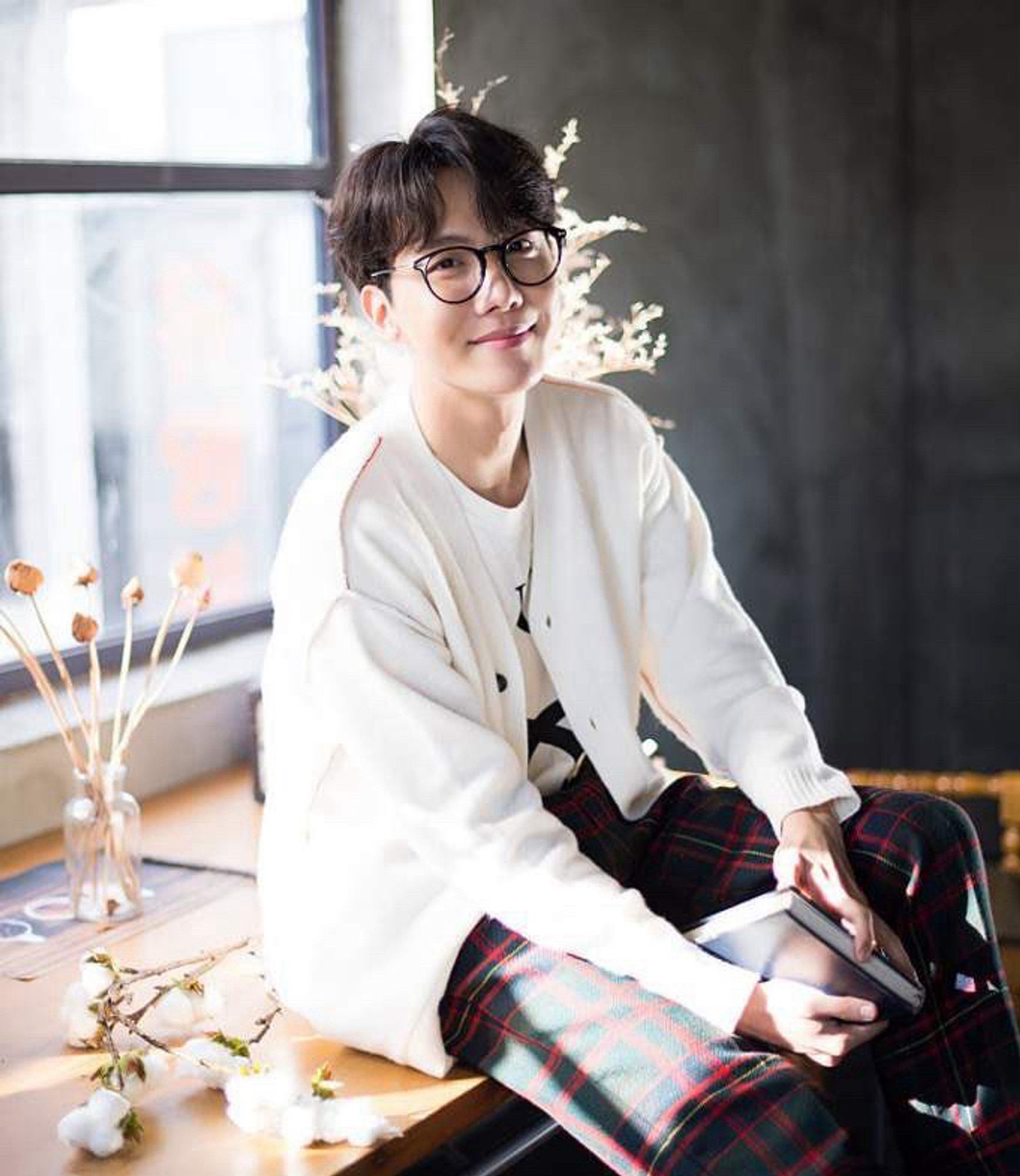 J-Hope of BTS in cream cardigan & glasses. He is sitting on a table beside a window holding a book