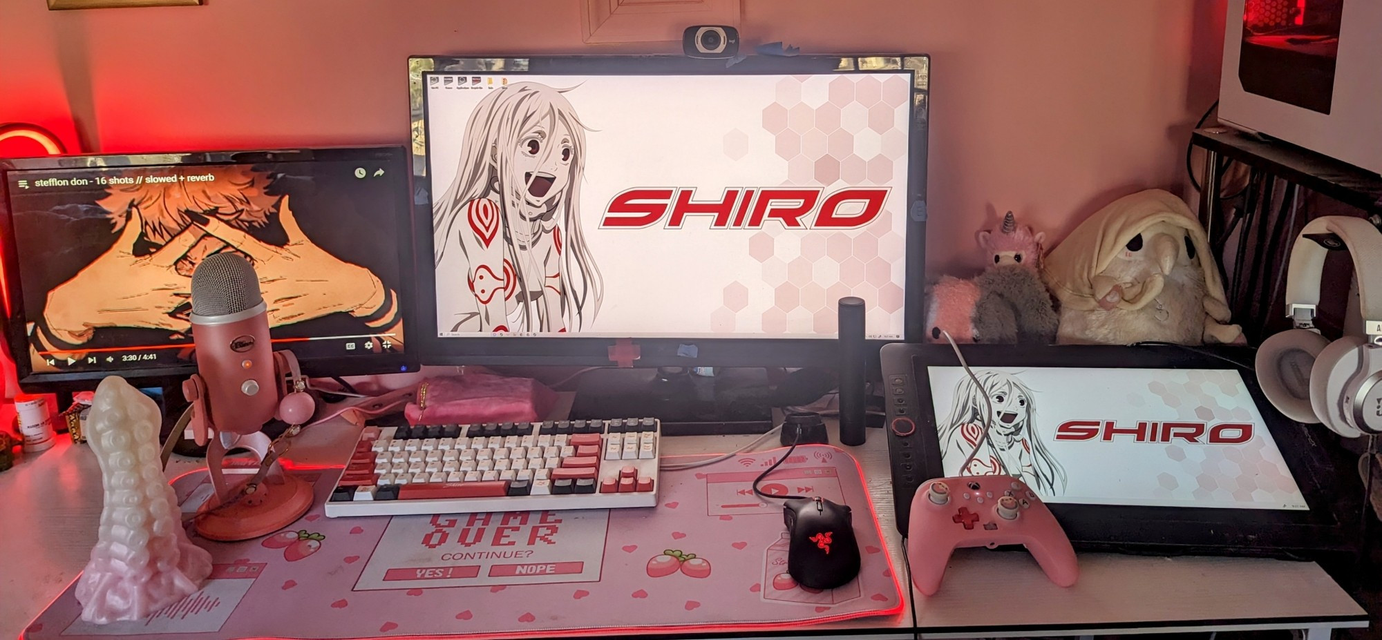 A photo of a gaming set up including a black, white, and red keyboard, a pink Xbox controller, three monitors, a pink mouse pad with strawberries that reads game over in the middle, a black razor mouse with the symbol glowing red a large yeti mic that has a pink ball gag hanging off of it, and a large tentacle dildo next to the mic for scale.