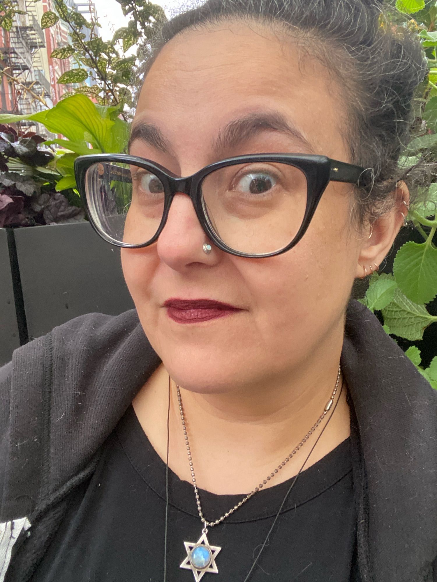 A white lady with curly brown and white hair piled on top of her head lifts her eyebrows at the camera. She has a nostril piercing and several ear piercings visible, as well as dark oxblood-stained lips. She is wearing a black dress and hoodie, with 2 necklaces, including a Star of David with a big moonstone at the center. There are fake plants behind her in the outdoor cabana thing that's really on Houston street amidst crazy traffic.