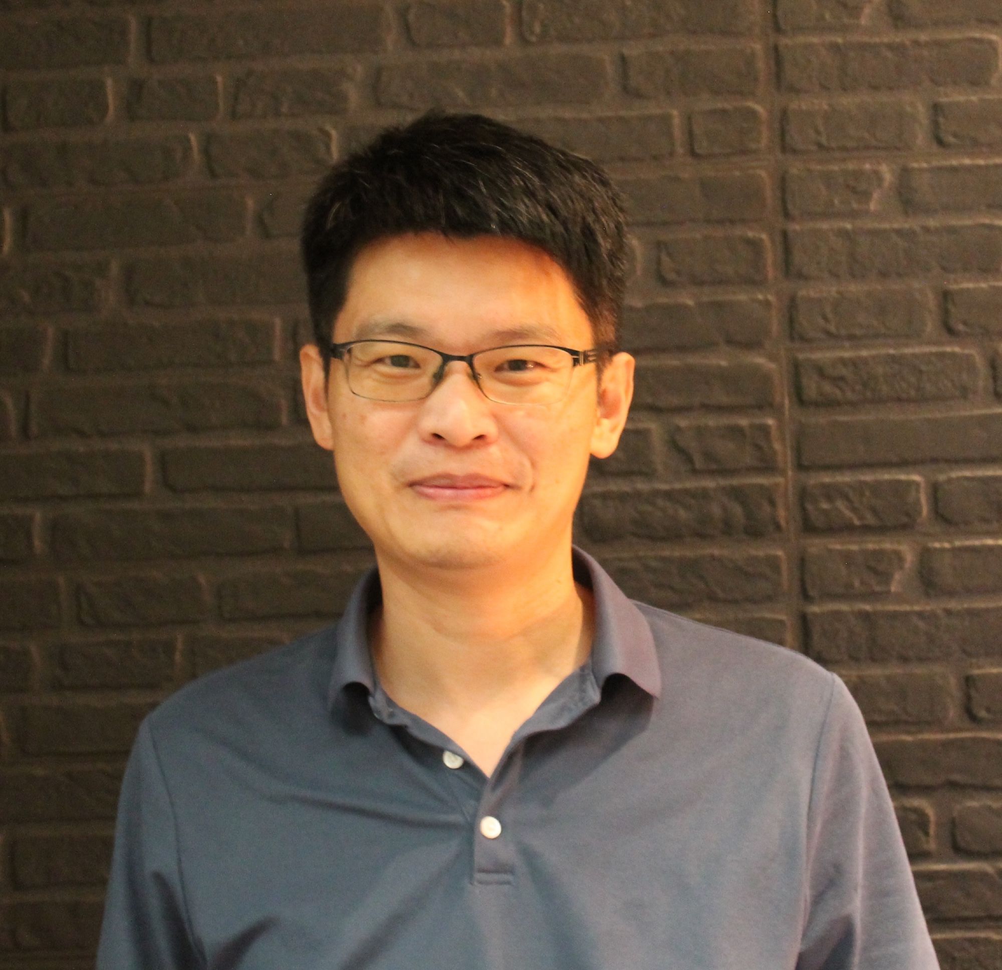 Headshot Kuo-Jung Lee (man with short black hair and glasses in gray polo shirt, standing against brick wall