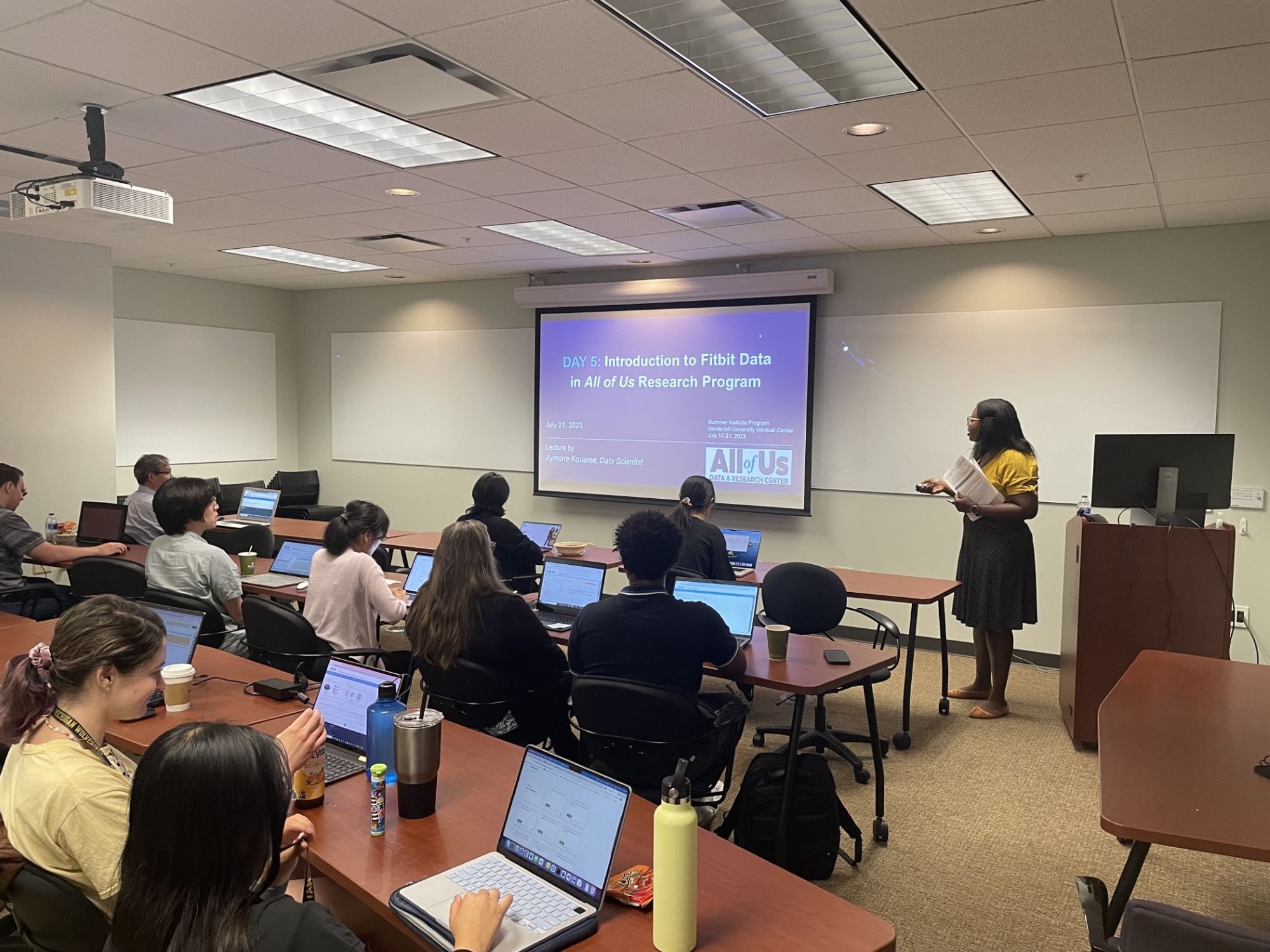 An African American woman in yellow blouse and black skirt stands next to a projected slide with the title "Day 5: Introduction to Fitbit Data in the All of Us Research Program." Three rows of students are visible; each one is sitting in front of a laptop.