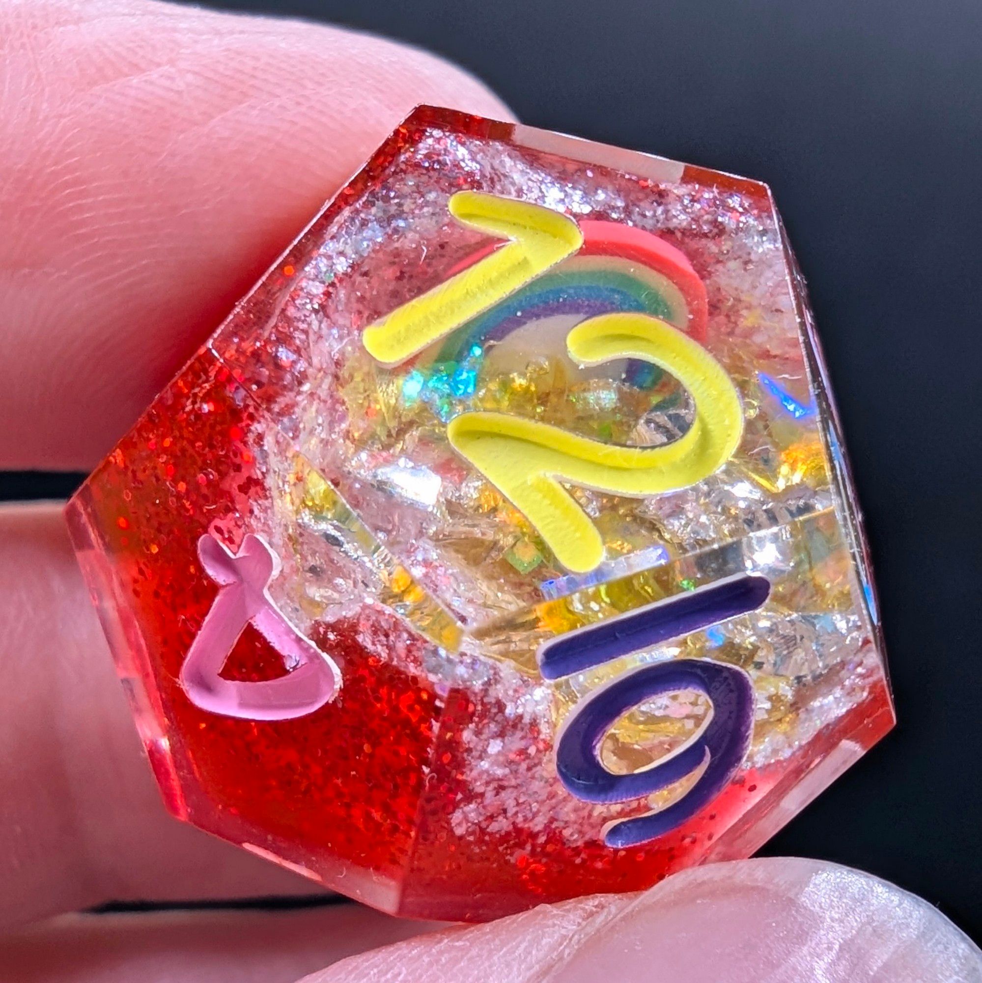 A close up of a d12 from a set of polyhedral dungeons and dragons dice. These are geode dice. The base is glittery ruby red, and the inside dung out part is painted a glittery white with yellow glitter flakes and a little rainbow inclusion.