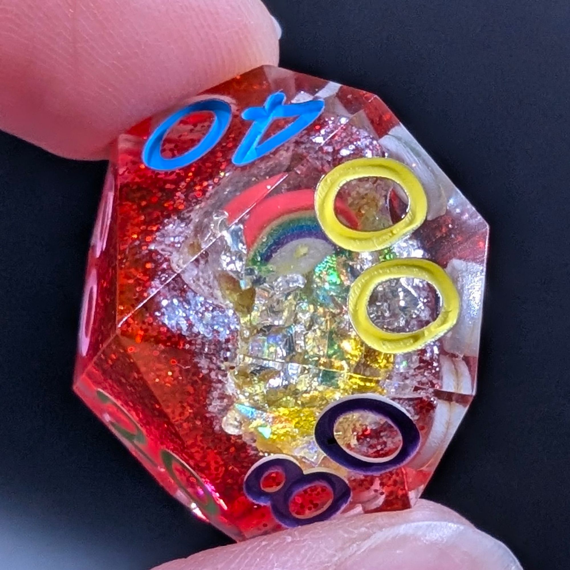 A close up of a d100 from a set of polyhedral dungeons and dragons dice. These are geode dice. The base is glittery ruby red, and the inside dung out part is painted a glittery white with yellow glitter flakes and a little rainbow inclusion.