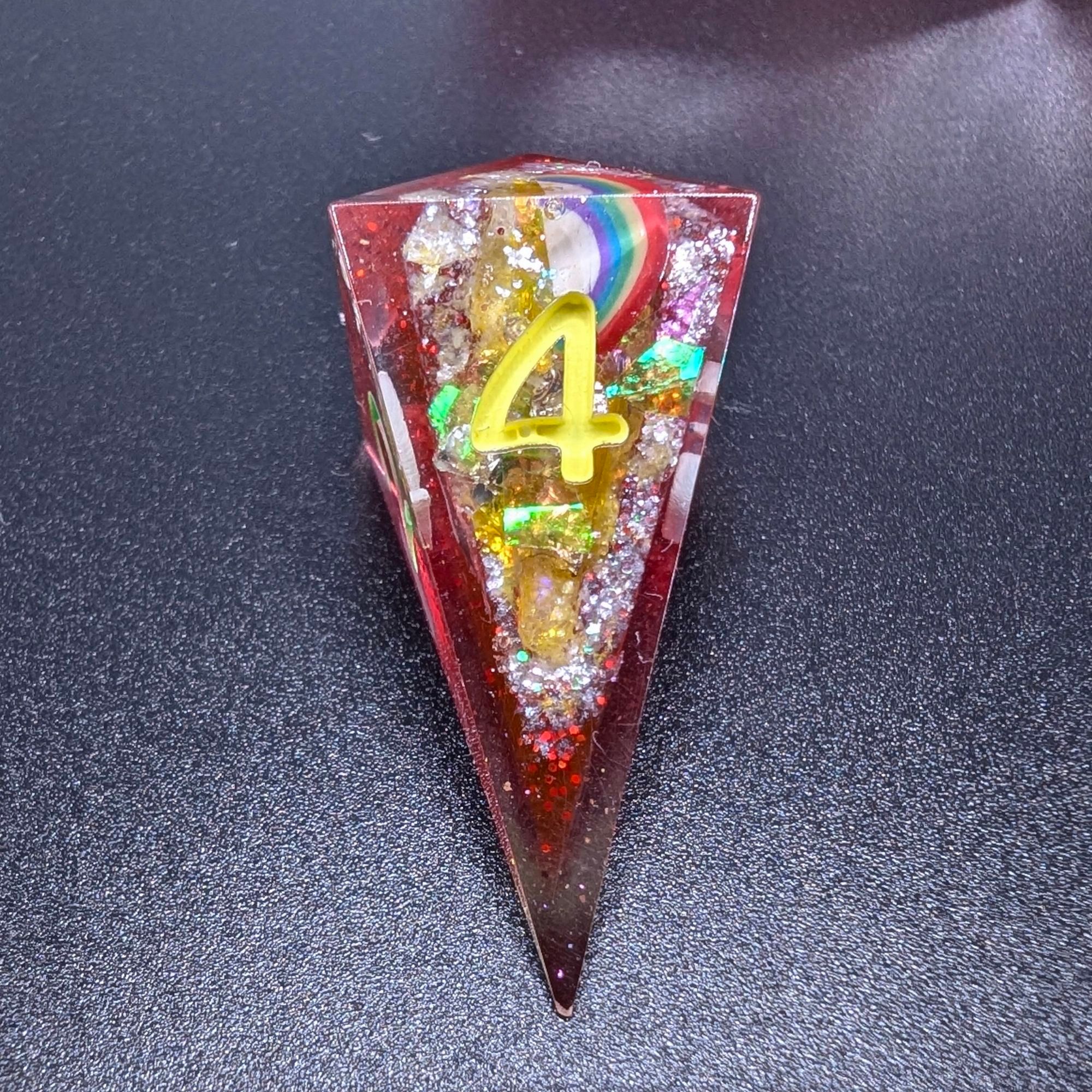 A close up of a shard d4 from a set of polyhedral dungeons and dragons dice. These are geode dice. The base is glittery ruby red, and the inside dung out part is painted a glittery white with yellow glitter flakes and a little rainbow inclusion.