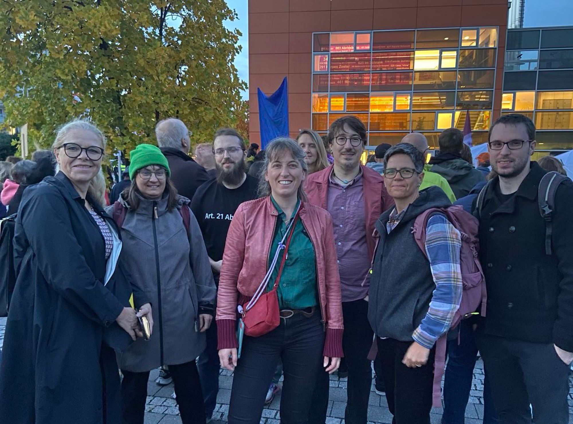 Eine Gruppe Menschen blickt in die Kamera. Im Hintergrund stehen einige Hundert Menschen vor dem Kulturhaus.