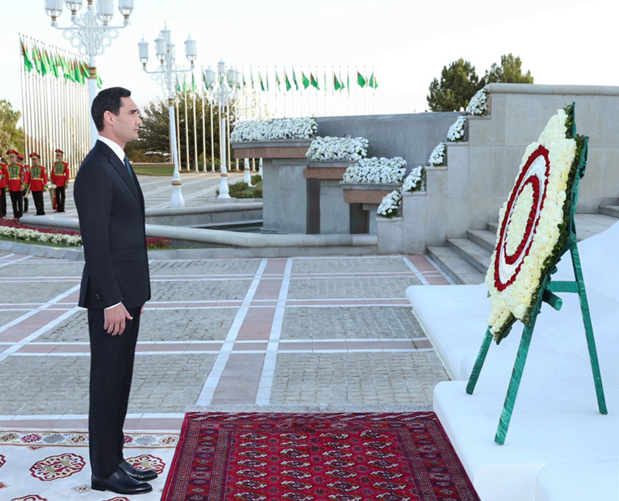 Serdar Berdimuhamedov by a wreath that looks like a dart board. There are carpets, outdoors.