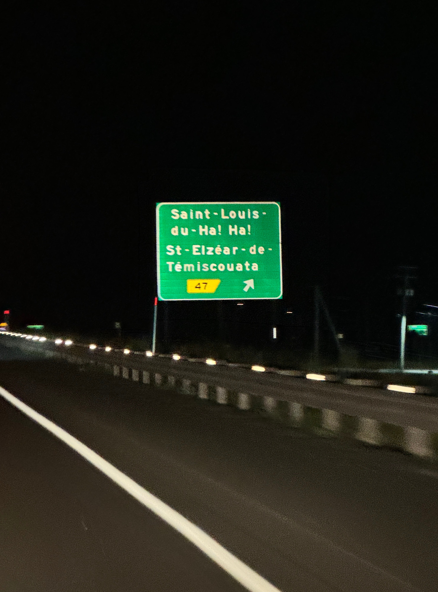 A highway road sign, on the side of the road, lit by headlights, reading “Saint-Louis-du-Ha! Ha!l.