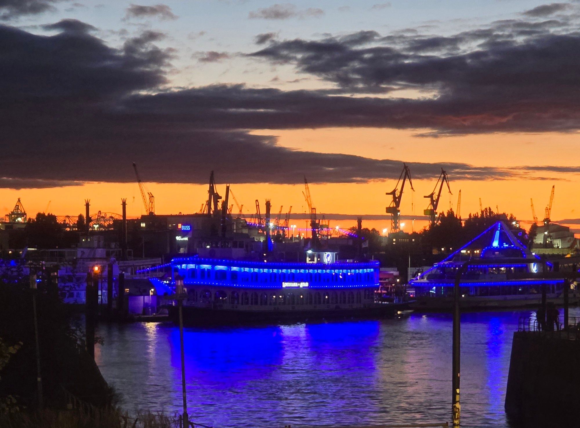 Blau beleuchtete Schiffe vor Hafenkränen und orangefarbenem Himmel mit dunklen Wolken