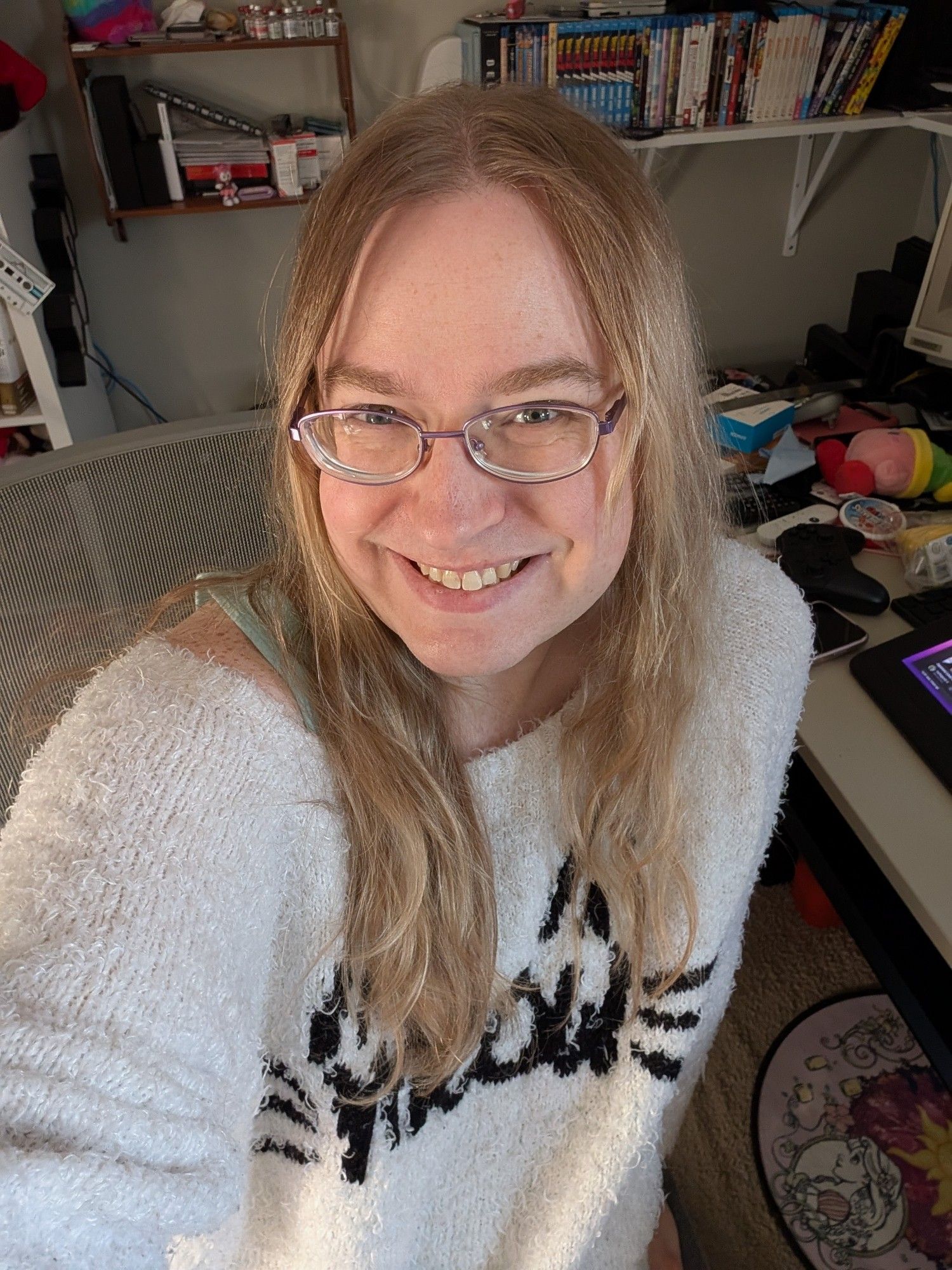 A selfie of a white transgender woman with long blonde hair and blue eyes wearing purple glasses and a white fuzzy sweater sitting in an office chair.