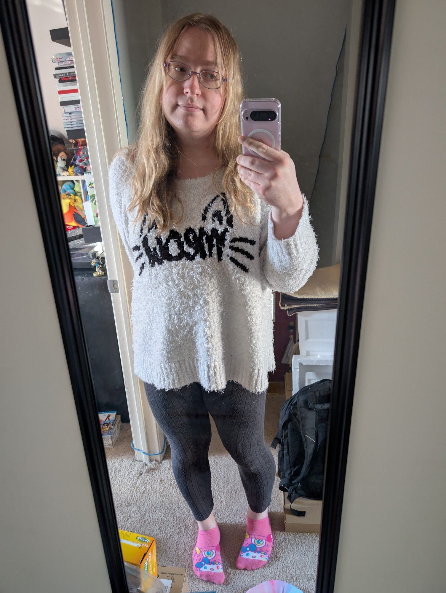 A white transgender woman with long blonde hair and blue eyes wearing purple glasses, a white fuzzy sweater with black text "meow" (mirrored) and cat ears above the "m" and "w", black leggings, and pink socks standing in front of a mirror.