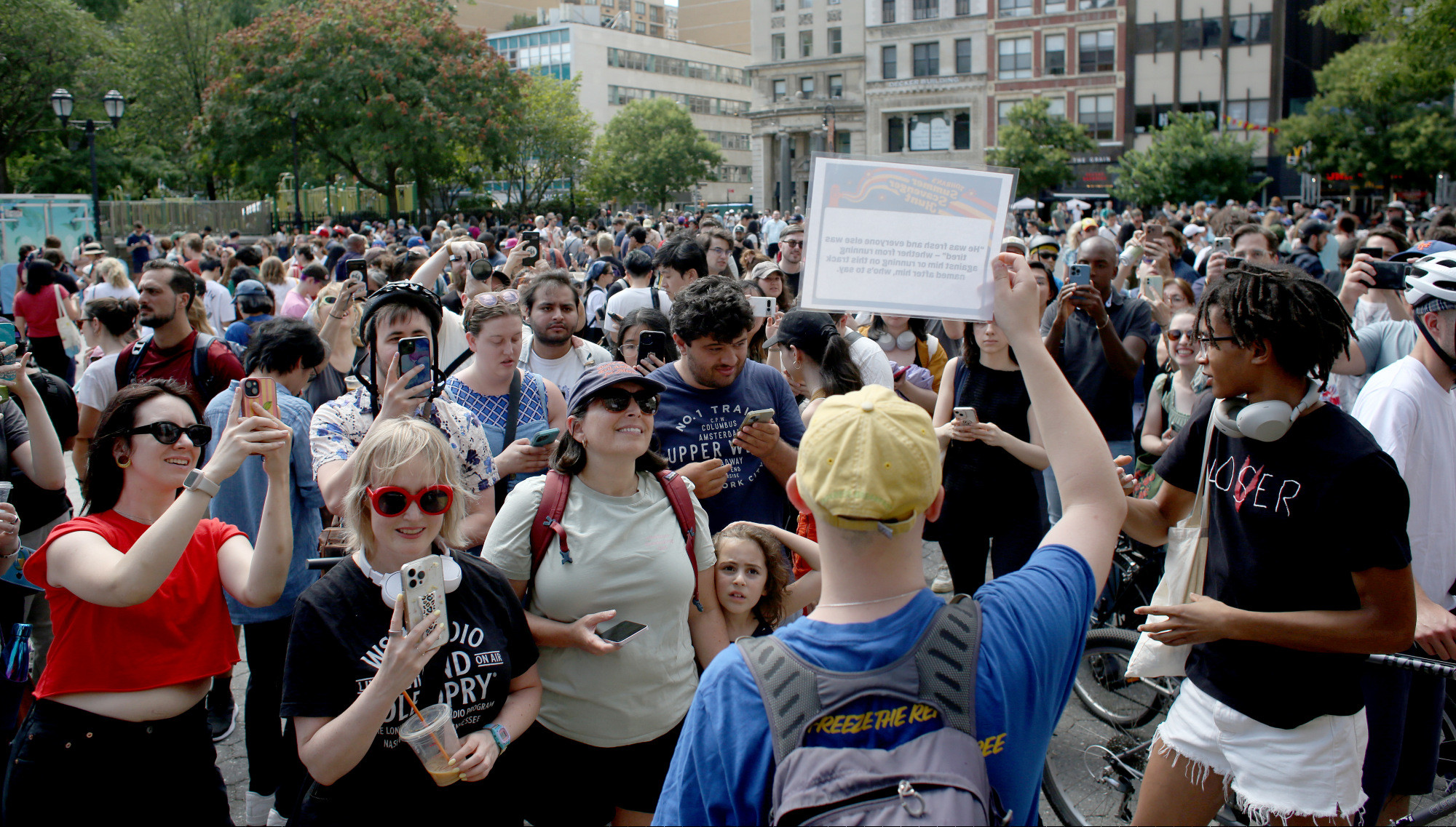 Mamdani scavenger hunt participants taking pictures of a card with the first clue that a campaign volunteer is holding up. Hundreds of people behind them in Union Square.