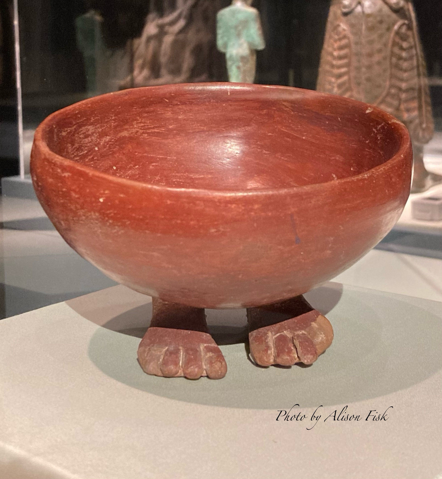 My photo shows a hemispherical red pottery bowl with supports in the shape of a pair of human feet on display at the Metropolitan Museum of Art in New York. Handmade from red Nile clay, it is slipped and polished to give a light surface sheen. The front of the bowl tips slightly forward as if offering its contents. Dimensions: diameter 13.2 cm x width 13.7 cm x depth 9.8 cm.