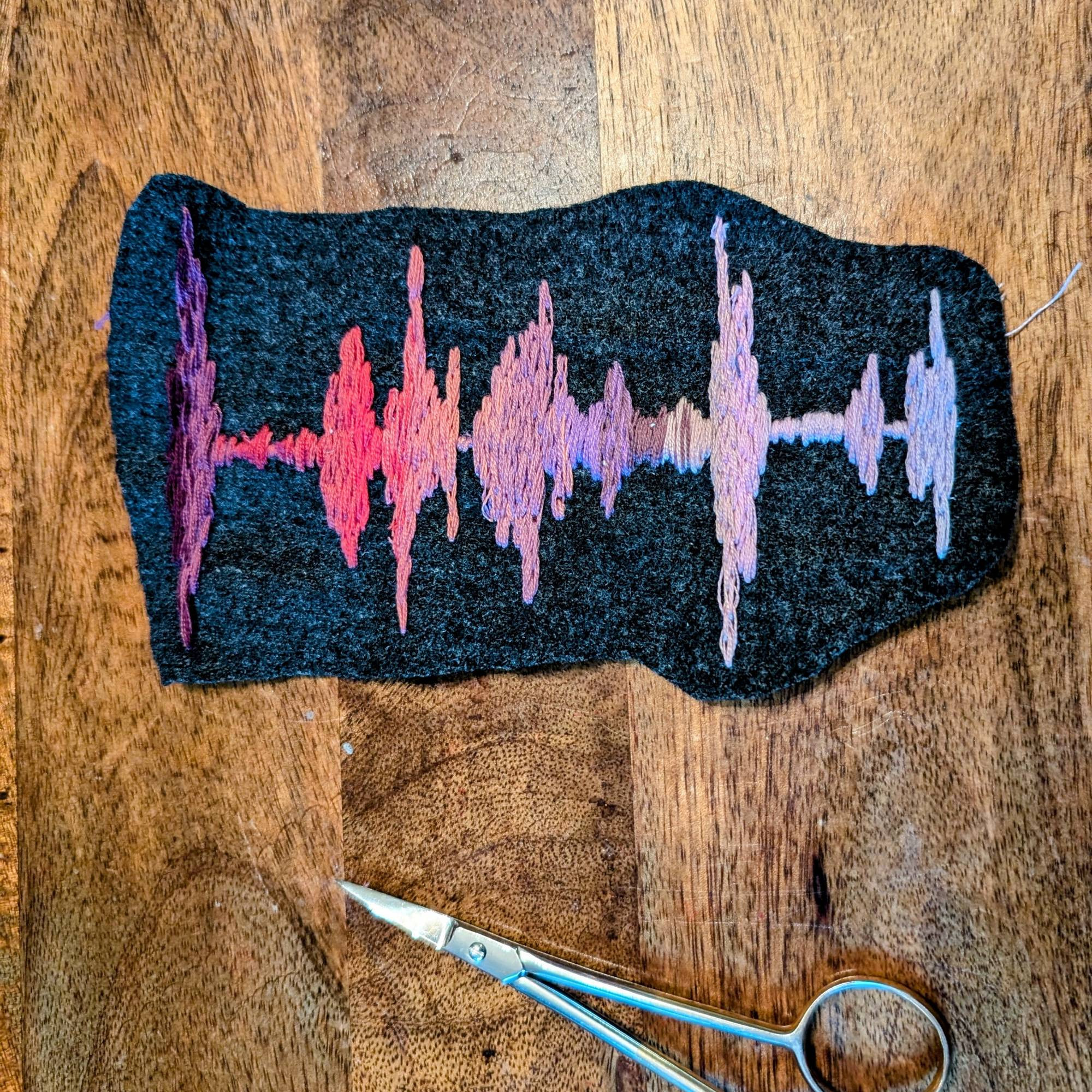 A purple-pink ombré embroidered sound wave, on a grey wool background , pictured with some embroidery scissors for scale 
