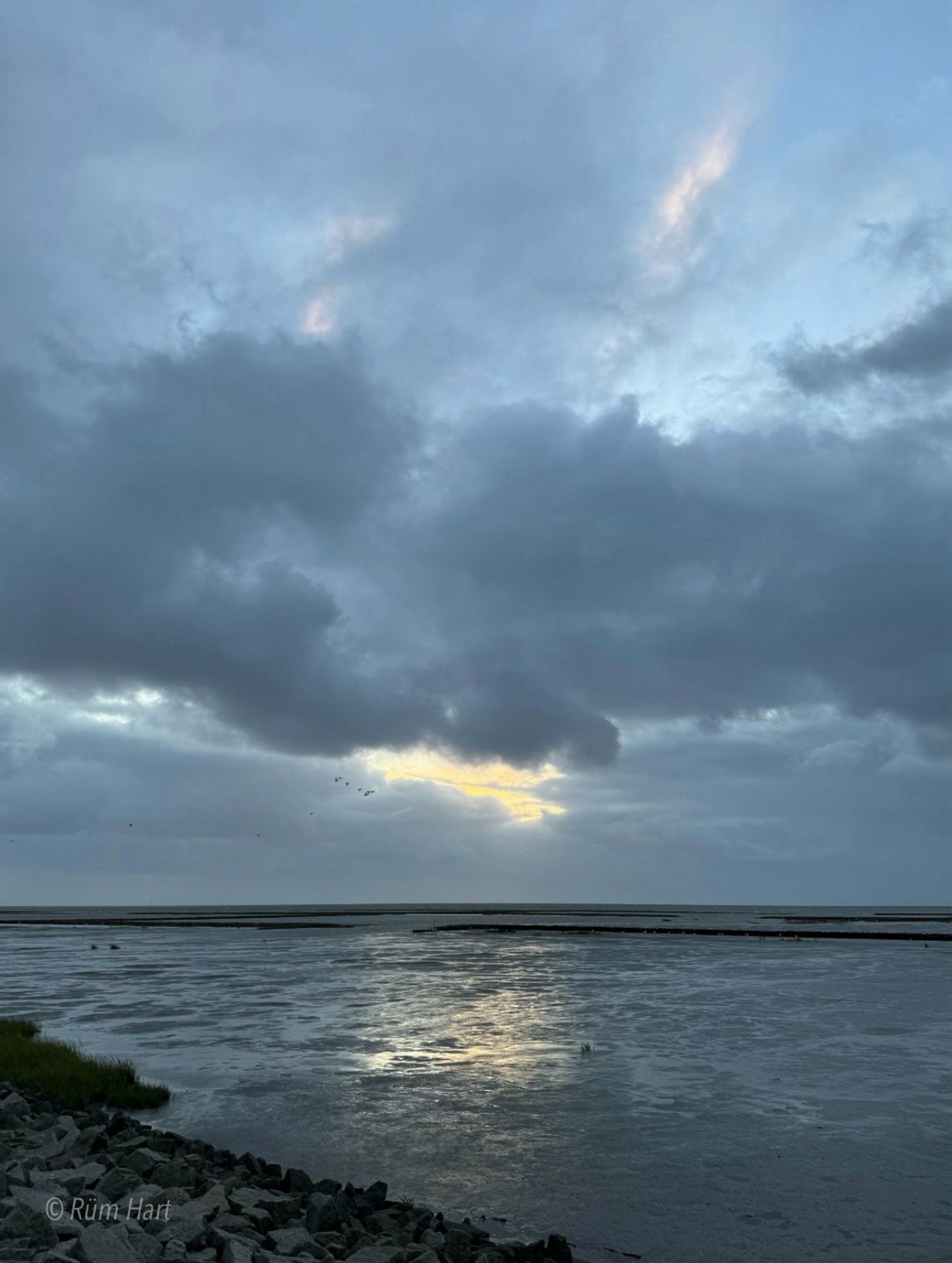 Blick auf das Watt. Ein bewölkter grauer Himmel, ungefähr in der Mitte, scheint die Sonne durch die Wolken. Oben sind zwei rosafarbene Stellen. 