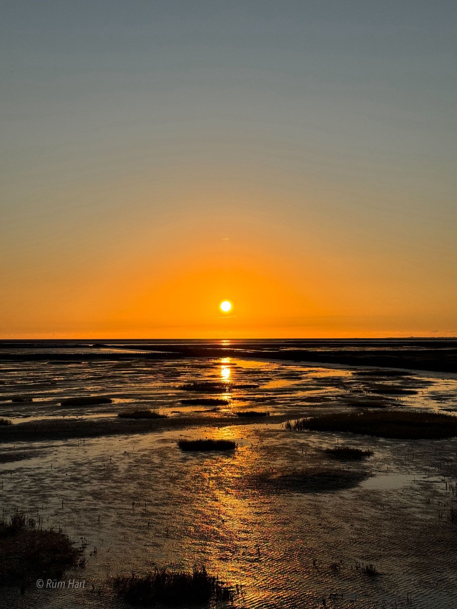 Ein orangefarbener Sonnenuntergang, der sich im Watt spiegelt an einem wolkenlosen blauen Himmel.