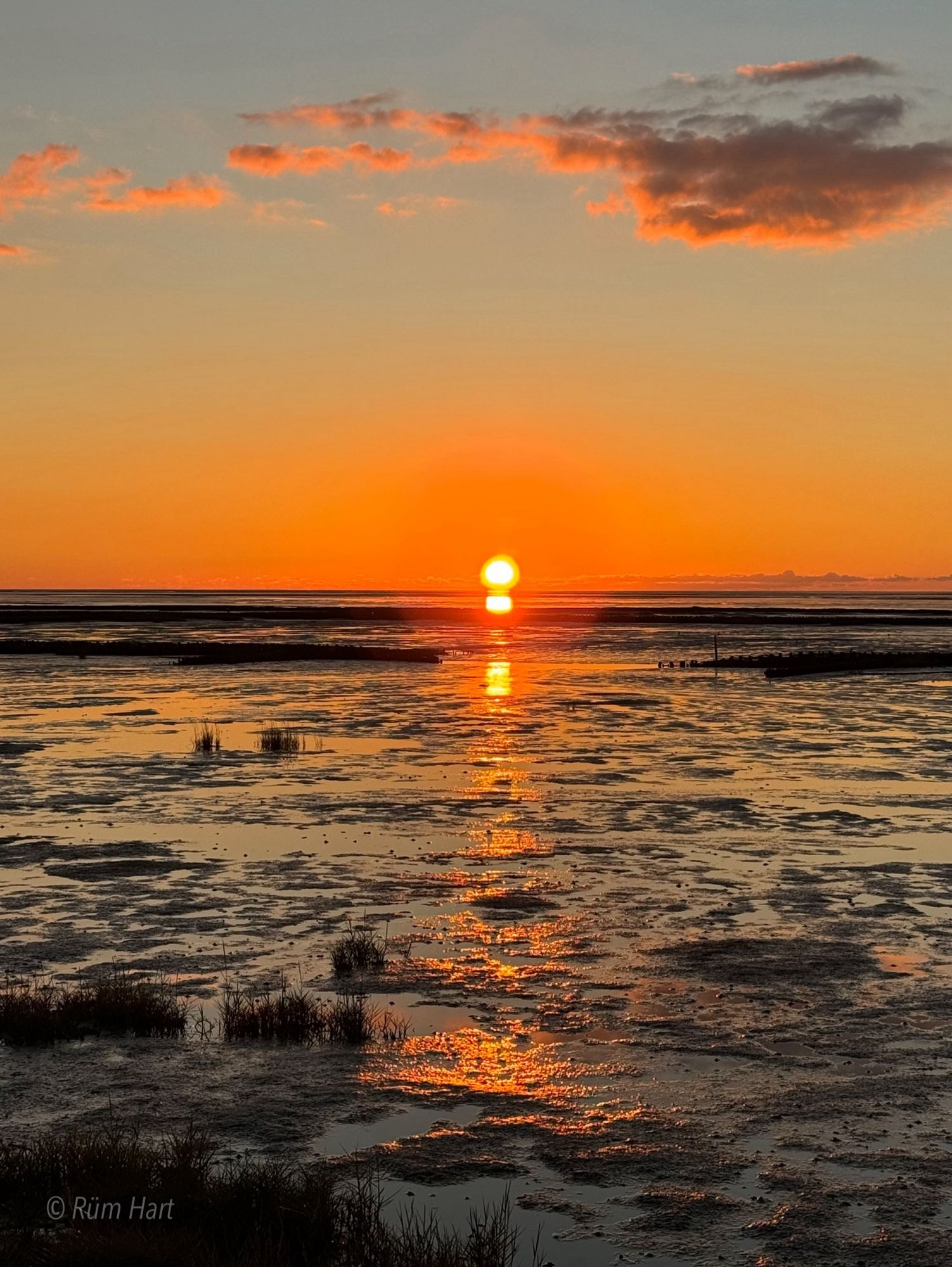 Ein orangefarbener Sonnenuntergang im Wattenmeer. Rosa Wolken sind am Himmel und die Sonne spiegelt sich teilweise im Watt.