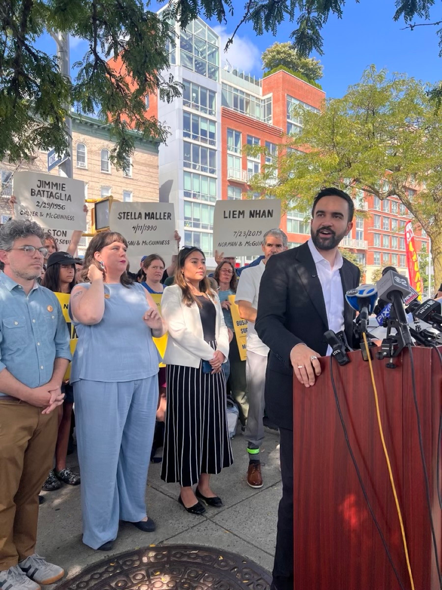 Zohran at a podium on a sidewalk. Behind him are Assemblymember Emily Gallagher, Councilmember Lincoln Restler and State Senator Kristen Gonzalez. Members of the Greenpoint community hold signs with the names of people who were killed on McGuinness Blvd. 