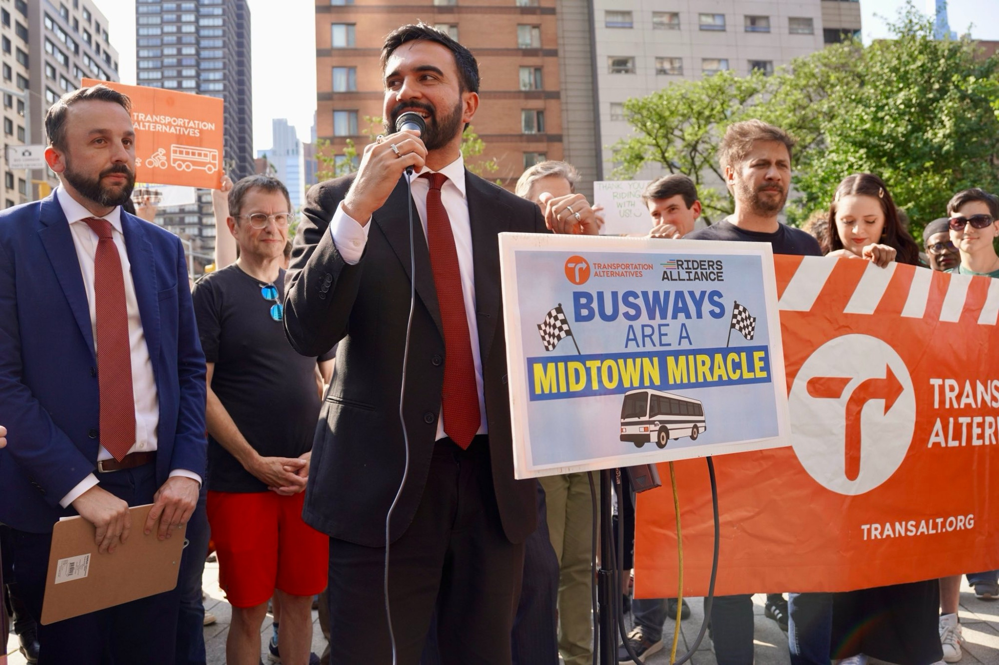 Zohran holding a microphone at a rally behind a podium and sign that says "Busways are a Midtown Miracle." Councilmember Keith Powers stands next to him, and advocates are holding signs and banners behind him.