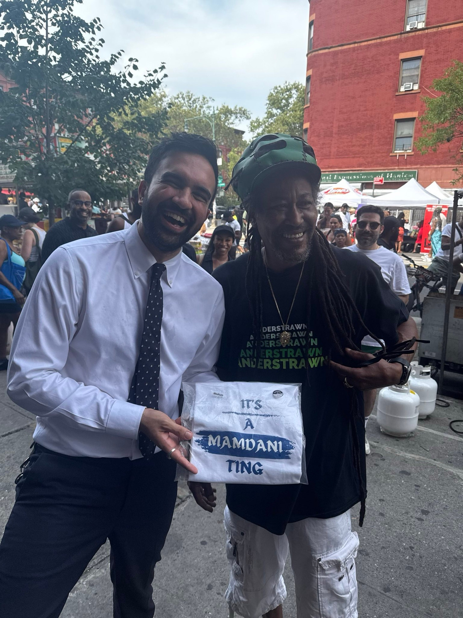 Zohran holding a shirt that says “It’s A Mamdani Ting” next to the man selling it. 