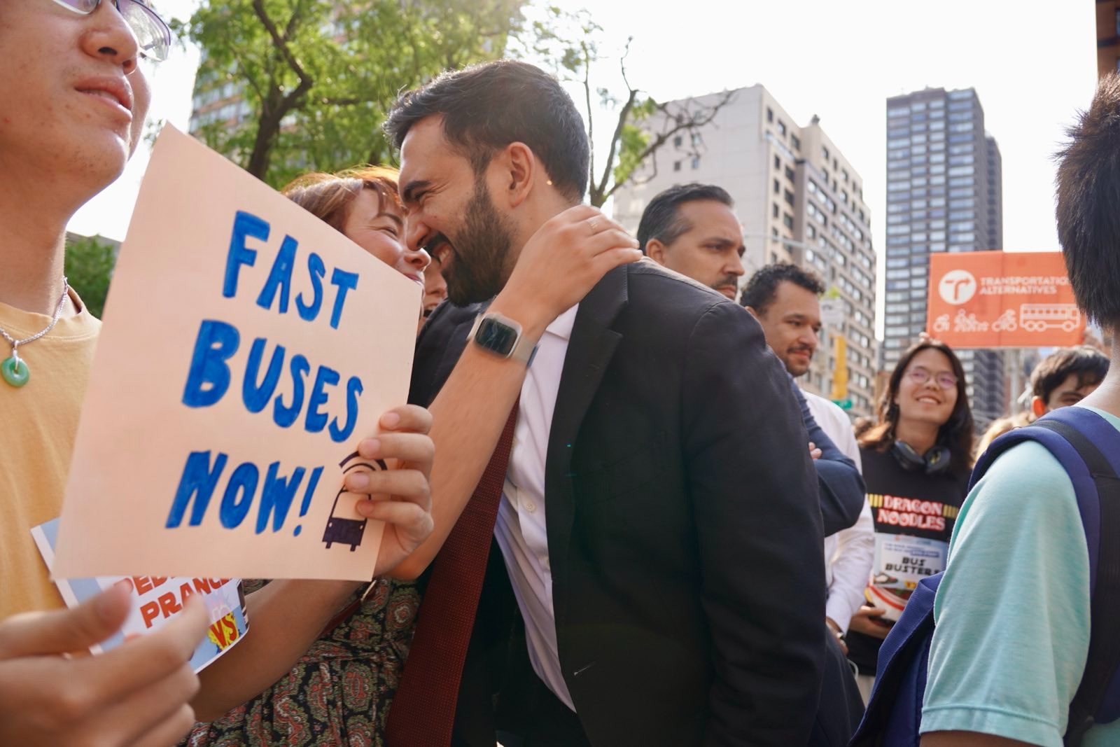 Zohran hugs an attendee at the bus race. Next to him someone is holding a sign that says "Fast Buses Now!" 