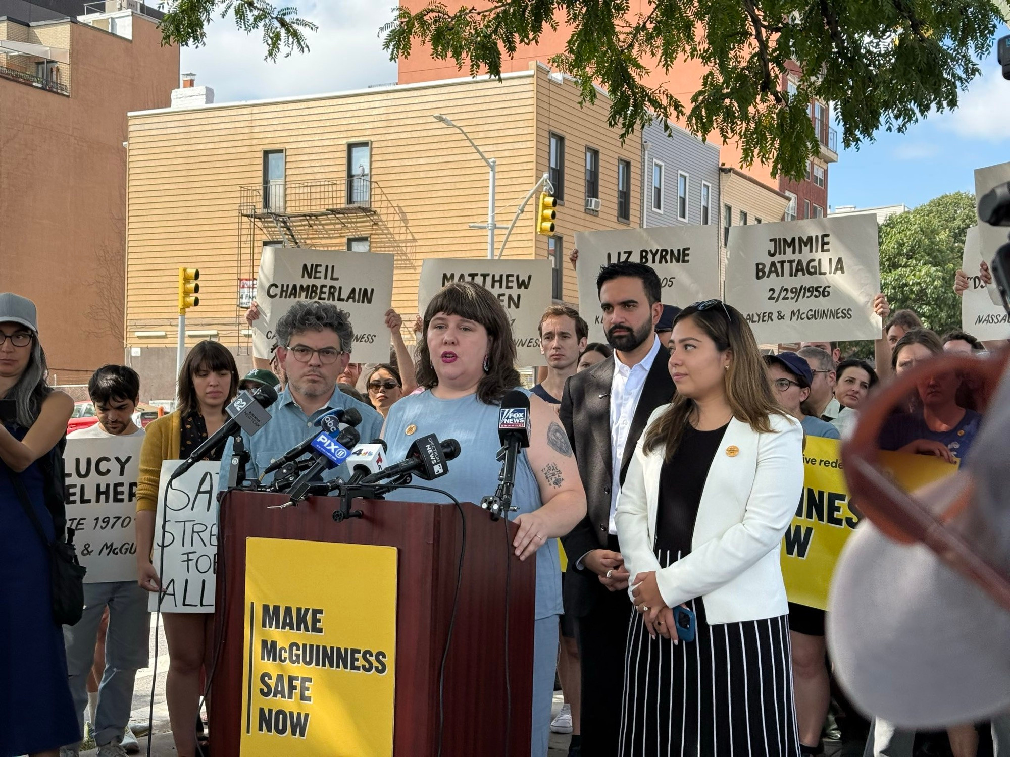 Assemblymember Emily Gallagher speaks at the podium, on the front of which is a sign that says “Make McGuinness Safe.”