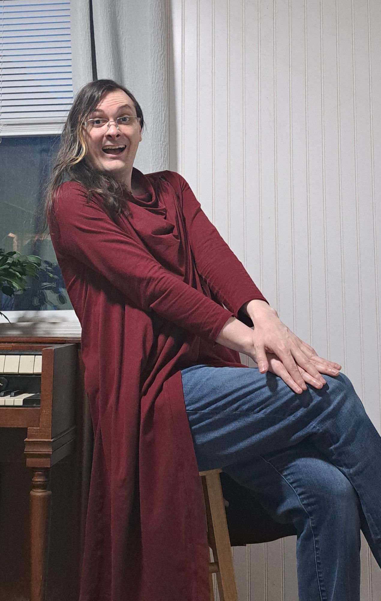 me facing toward the camera, sitting on a stool with my legs crossed to the side, hands overlapping on the knees, leaning back slightly toward an electric organ, and I'm smiling very excitedly with my mouth wide open. I'm wearing a long red cowl-neck top and glasses. my hair is a little past shoulder-length and is dark brown with a long streak that's been bleached blonde.