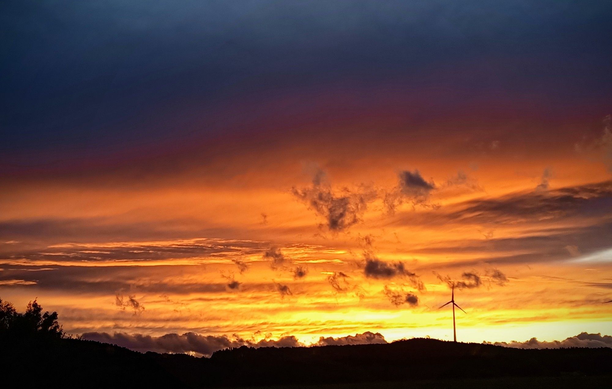 Schmale schwarze Silhouette mit einzelnem Windrad.Der Himmel nun hell über der Silhouette,dann gelb,feurig rot orange und darüber noch dunklere Wolken die ,die nahende Dunkelheit ankündigen