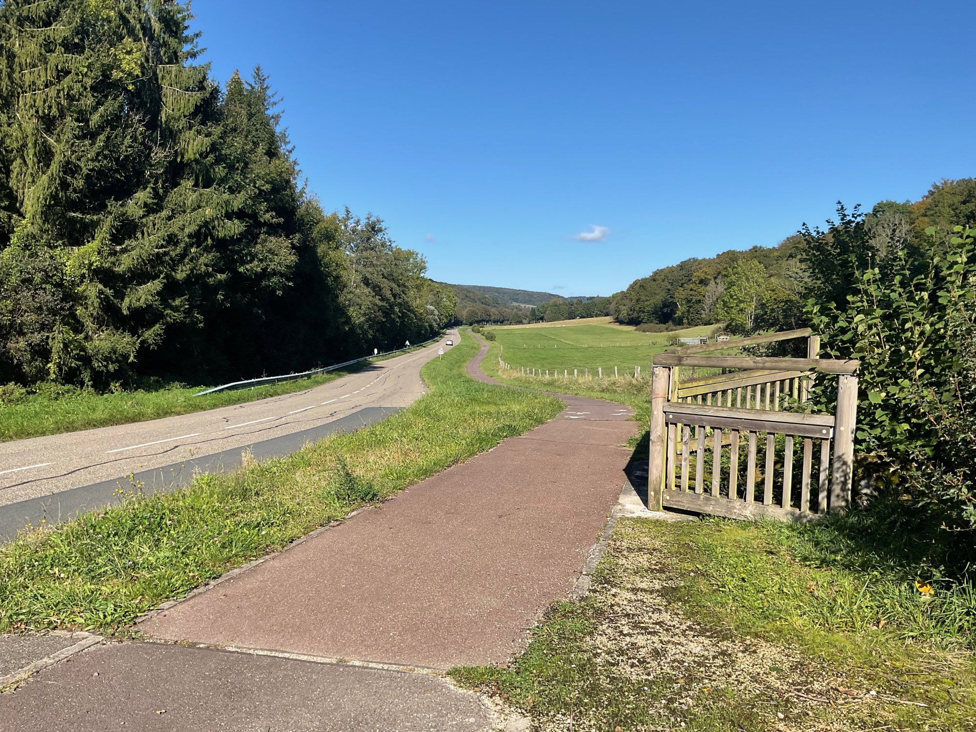 Foto, geschossen von einem Gehweg rechts neben einer Straße. Rechts, links und in der Ferne sind Wälder zu sehen. Rechts neben dem Gehweg befindet sich ein Holzzaun, der die (nicht zu sehende) Einmündung eines Bachlaufs unterhalb der Straße umschließt. Der Himmel ist strahlend blau, nur ganz in der Ferne sind drei winzigkleine Wölkchen zu sehen.