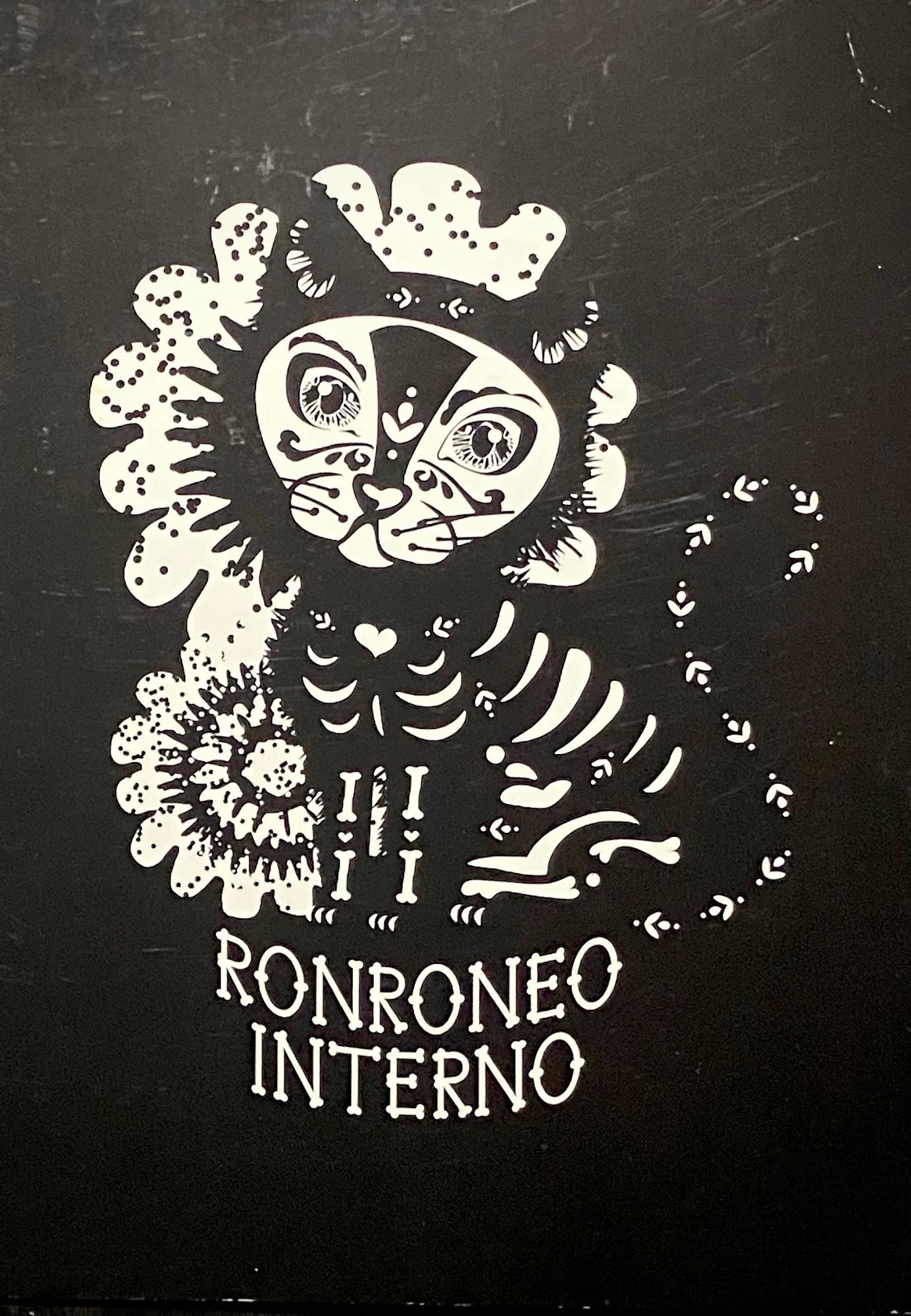 Postcard with a cat styled in Día de Muertos fashion with a visible skeleton, face make-up and floral detailing. The card is black and white, with the caption RONRONEO INTERNO