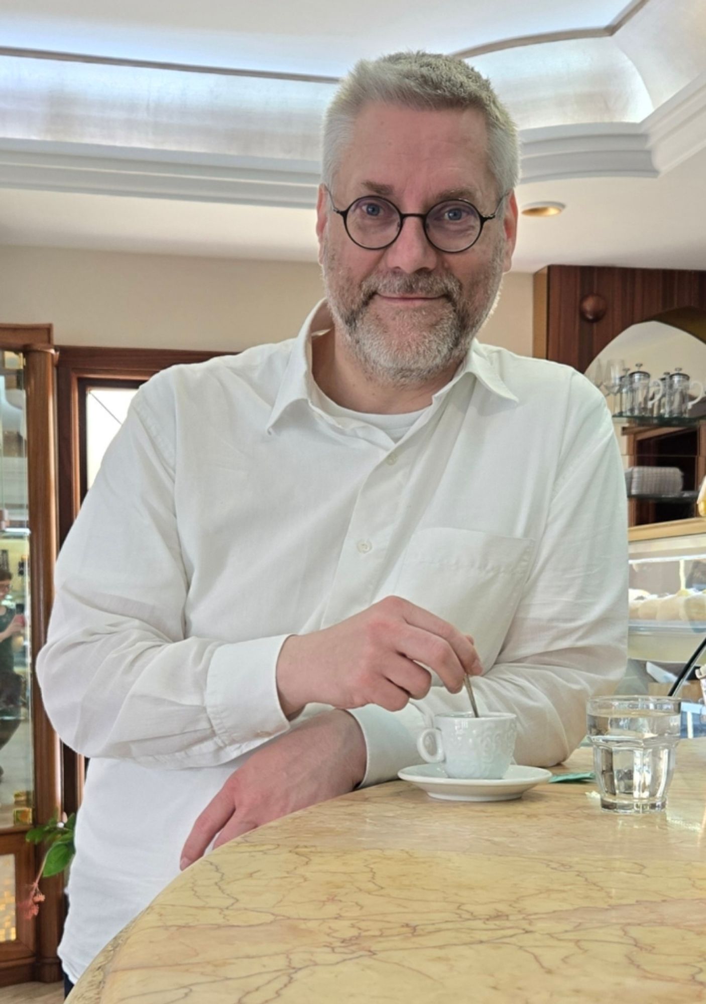 Freundlich schauender Bart- und Brillenträger mit grauen Haaren und weißem Hemd steht am Tresen und rührt in seinem Espresso.