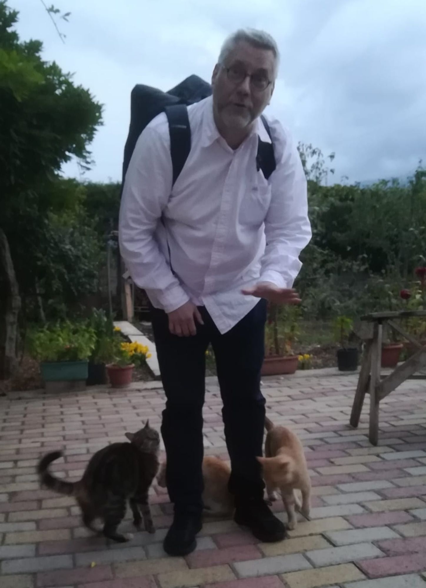 Ein grauhaariger Mann mit weißem Hemd und Jeans samt Rucksack. Und um die Beine drücken sich die drei Katzen.