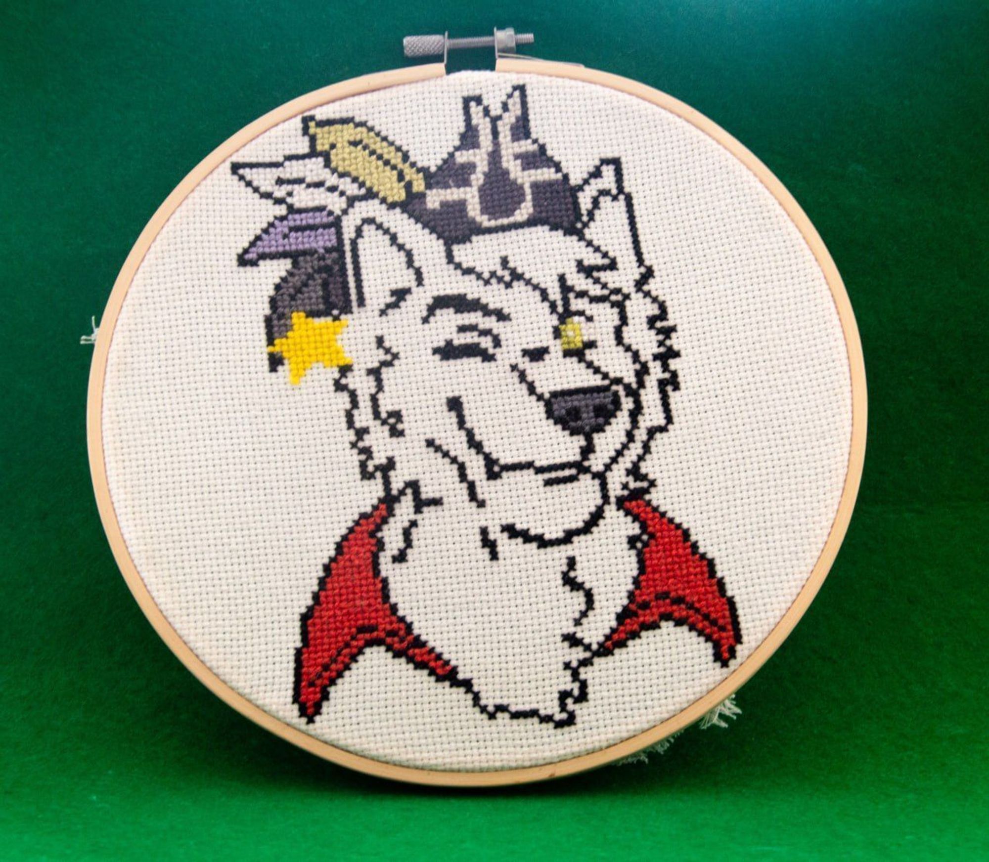 A cross-stich anthro wolf on a linen and wooden frame. The wolf is partially colored, dressed as a pirate with a pirate tricorn hat adorned with colored feathers, and winking one of their yellow eyes at the same time they smirk