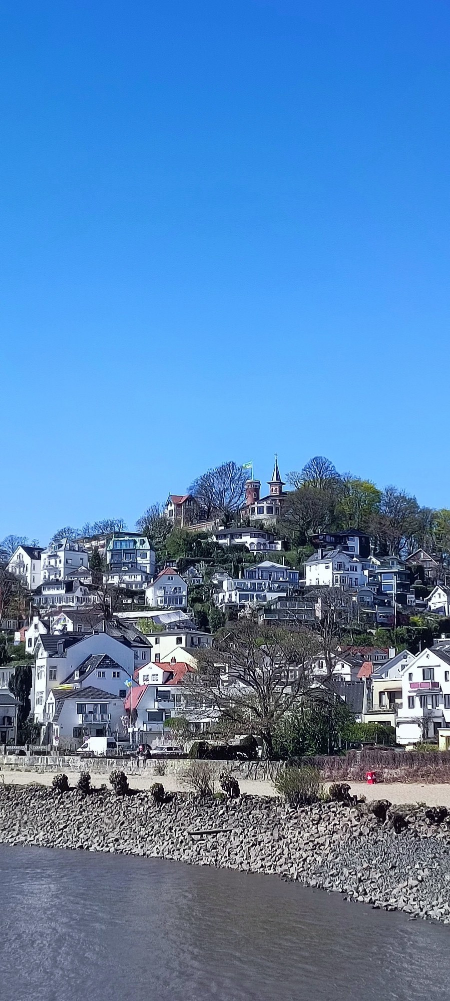 Blankenese von einer Fähre aus mit pituresken Häusschen die sich Seite an Seite auf einen eigentlich zu kleinen Hügel breit machen.Im Hintergrund der blaue Himmel eines tollen Tages.