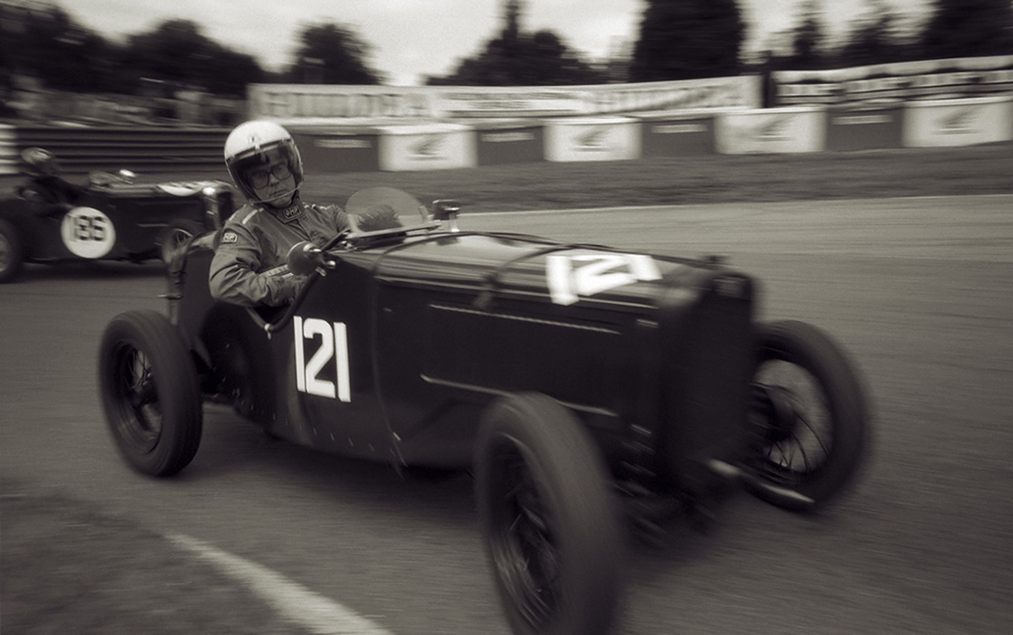 A black and white photo of a pre-war Austin Seven entering the hairpin at Mallory Park. The driver is looking down and making eye contact with the camera, which is held very low at the apex of the corner.