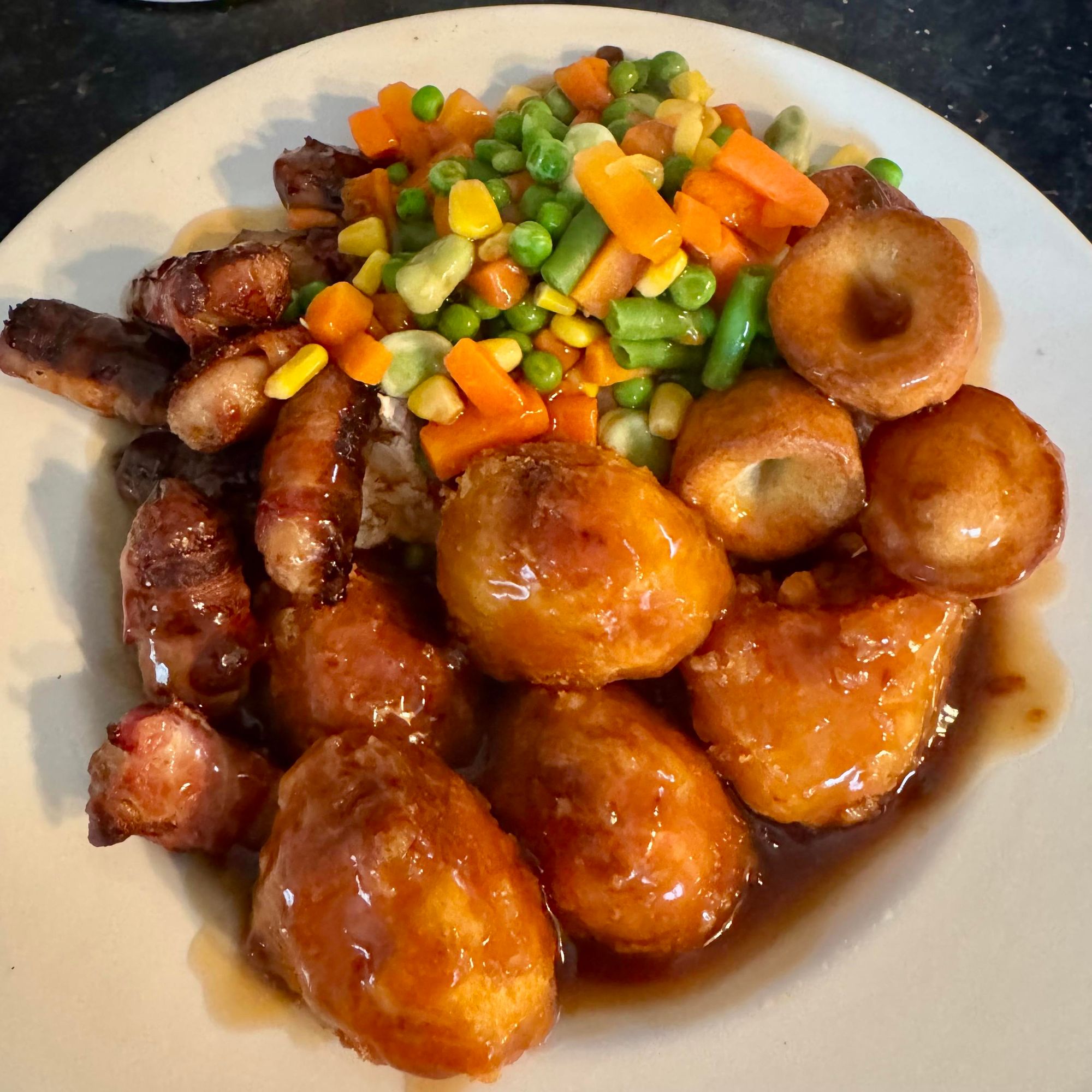 A plate of food featuring roast potatoes, Yorkshire puddings assorted vegetables (carrots, peas, sweetcorn, green beans & broad beans), kilted sausages and roast chicken, all drizzled with gravy.