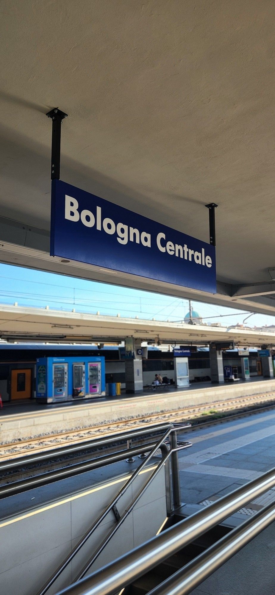 A imagem mostra uma placa azul com o texto branco 'Bologna Centrale', pendurada no teto de uma plataforma de estação de trem. Ao fundo, podem ser vistos os trilhos e outra plataforma, com máquinas de venda automática e algumas pessoas esperando. A estação parece limpa e organizada, com corrimãos de metal brilhante em primeiro plano.