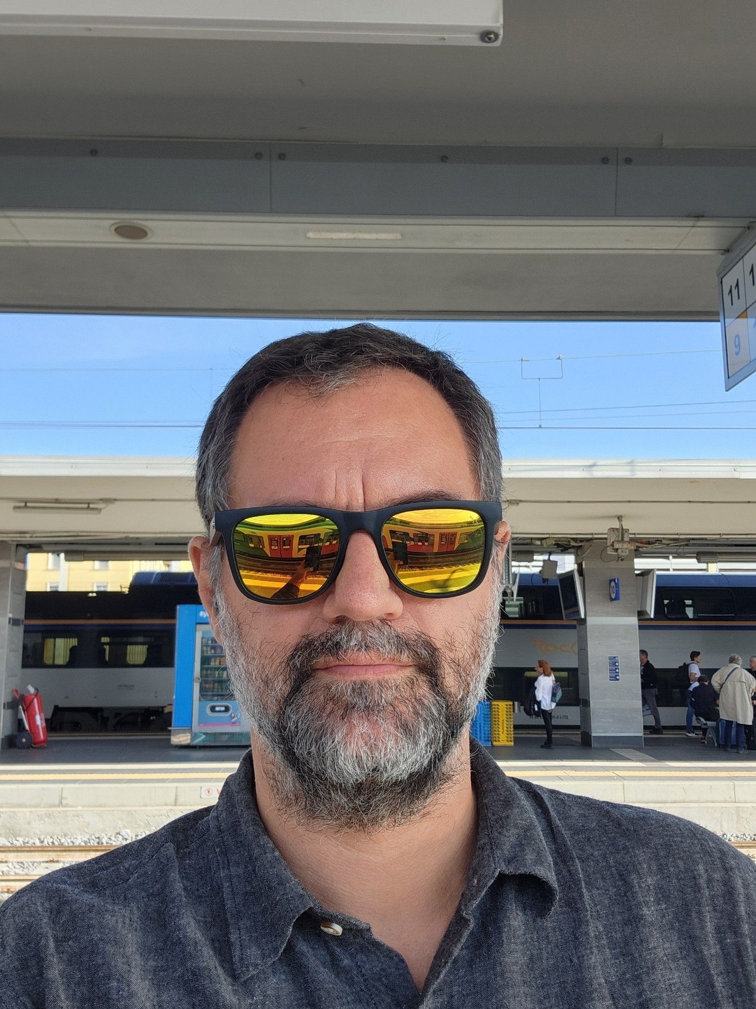 Na imagem, Érico Andrei está em um ambiente de estação de trem, usando uma camisa cinza escura e óculos de sol espelhados com lentes amarelas. Ao fundo, vê-se uma plataforma de trem com algumas pessoas, vagões estacionados, e um céu claro. O reflexo das lentes dos óculos de sol de Érico mostra parte da estação e vagões de trem.