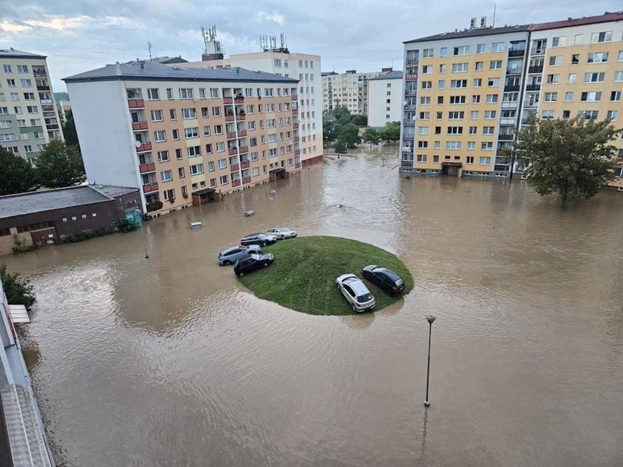 The photo shows a flooded area with several high-rise buildings. In the middle is a grass-covered hill surrounded by the floodwaters. Five cars are parked on it.