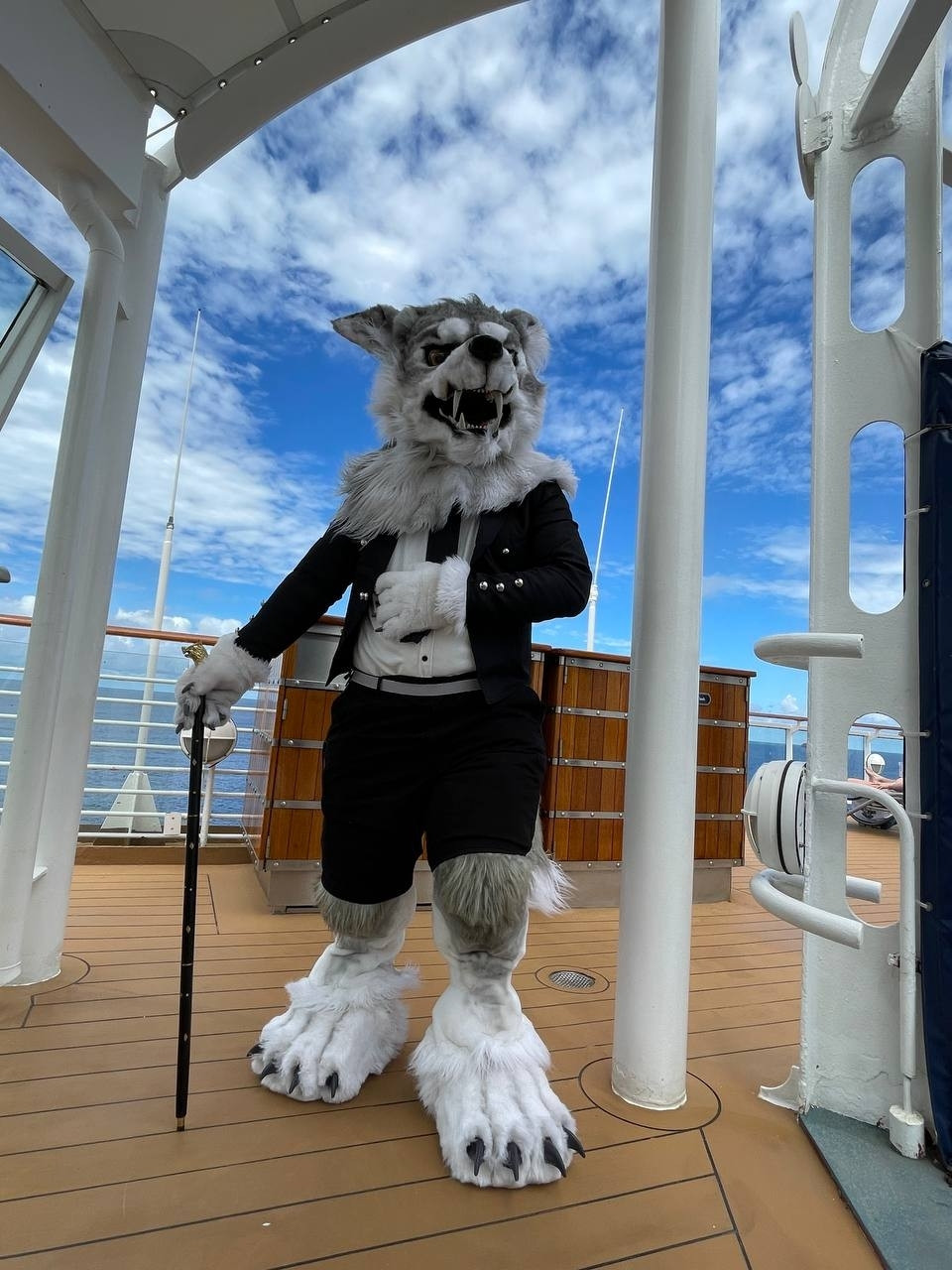 Cray the Werewolf is wearing his fancy tuxedo and holding a wolf-headed cane while standing by the guard rail of a cruise ship under a blue sky.