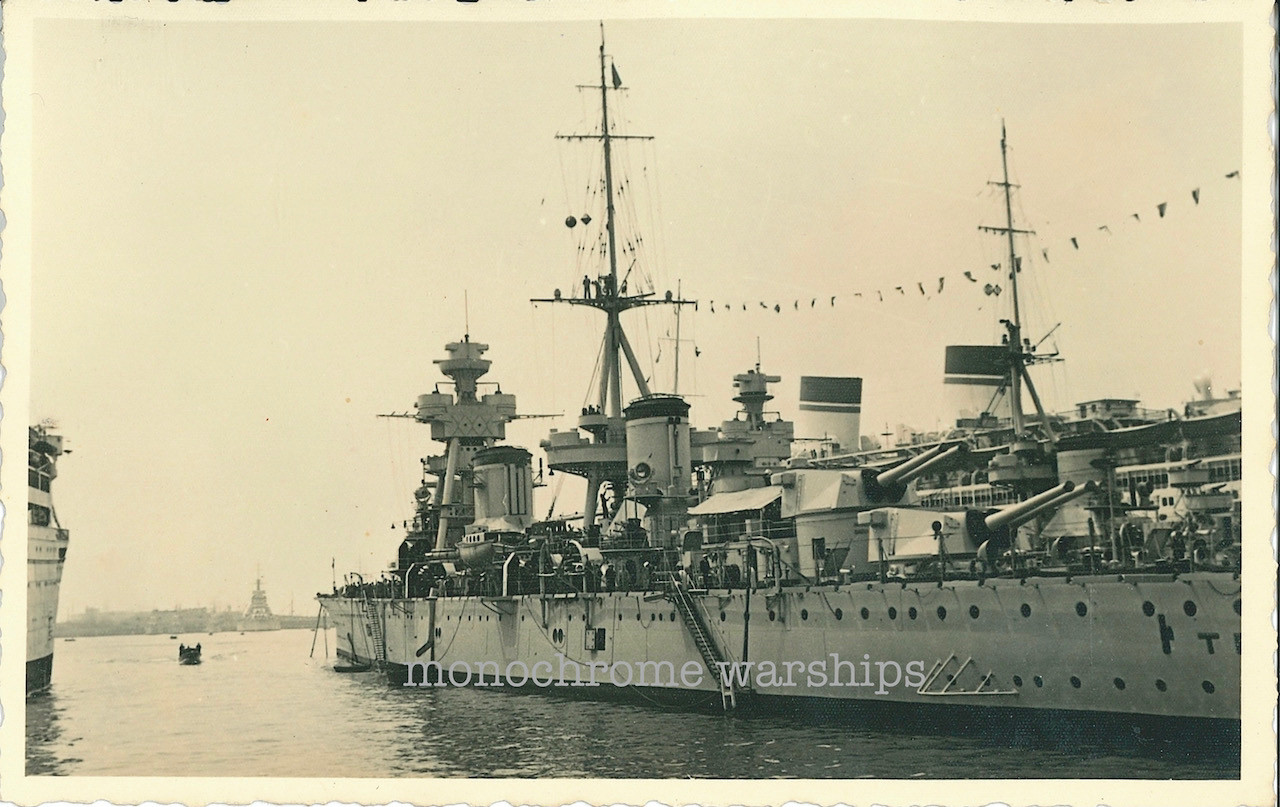 A photo of the two Trento-class heavy cruisers looking in 3/4 profile from astern. Behind are the distinctive striped funnels of the liner Conte di Savoia. In the distant background can be seen the shape of a Conte di Cavour-class battleship.