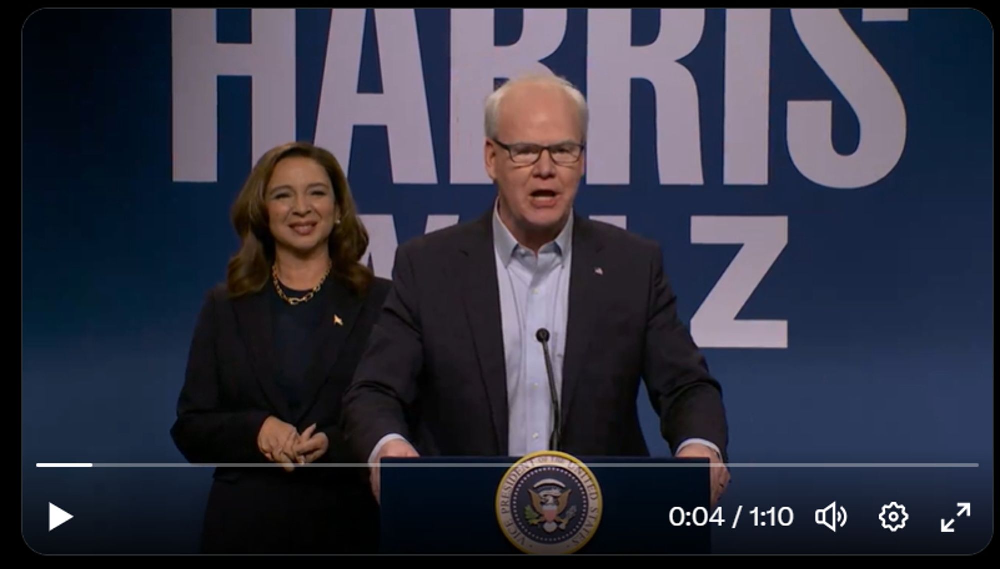 A screenshot of Maya Rudolph as Kamala Harris standing behind Jim Gaffigan playing Tim Walz standing at a faux presidential lectern speaking to the audience on Saturday Night Live