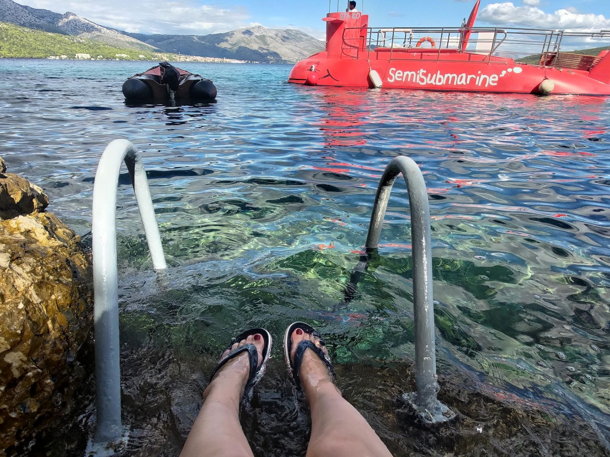 Women's feet in Crocs on the sea, at the Mediterranean, with a weird semi-sub in the background, and more importantly, the location where there best Croatian wines are made a bit further in the background